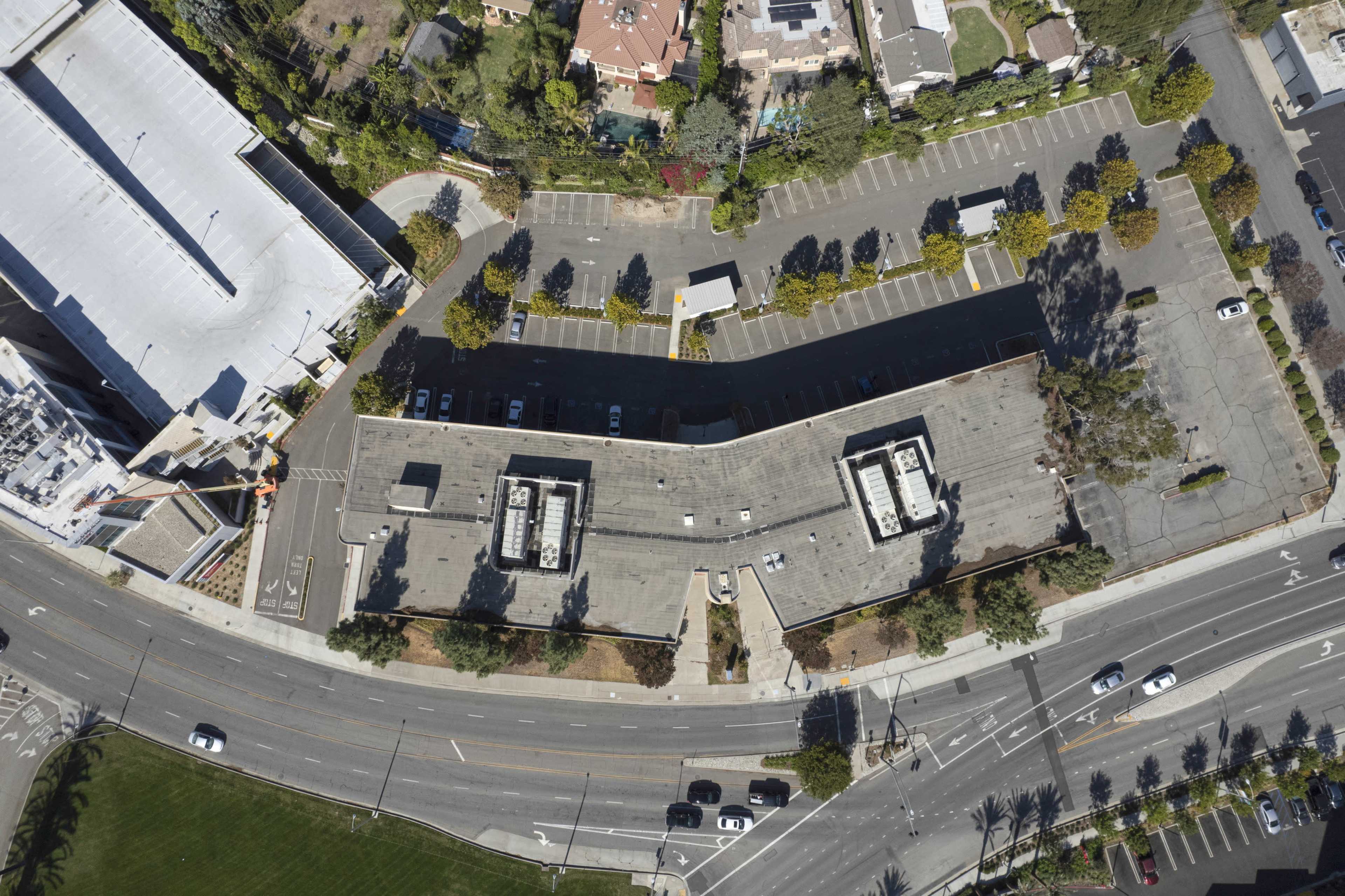 This aerial view shows a commercial building with a large parking lot adjacent to a busy road, surrounded by trees and other structures.
