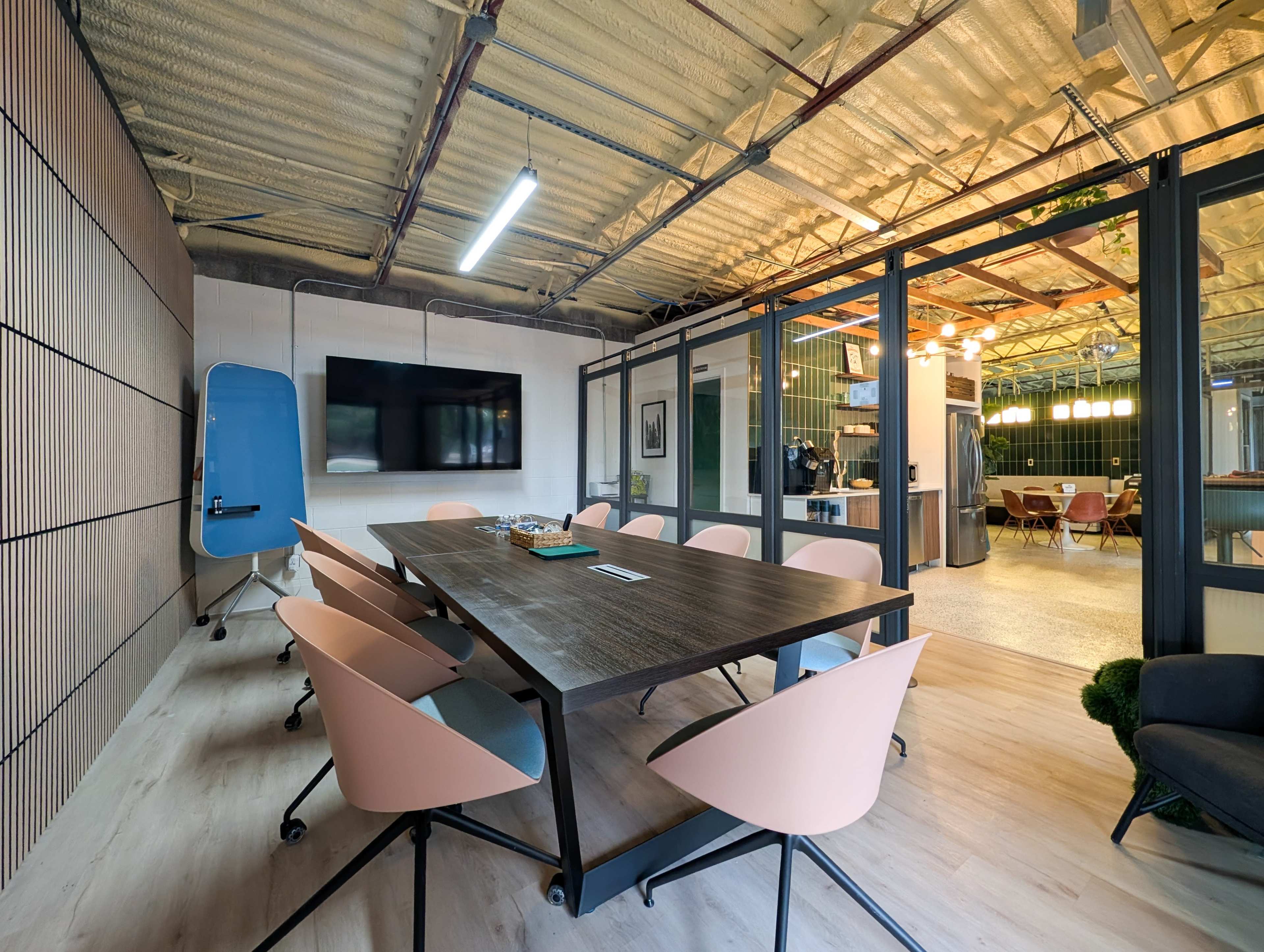 A modern conference room features a large wooden table surrounded by eight chairs, with a wall-mounted screen and a glass partition view into a brightly lit area.