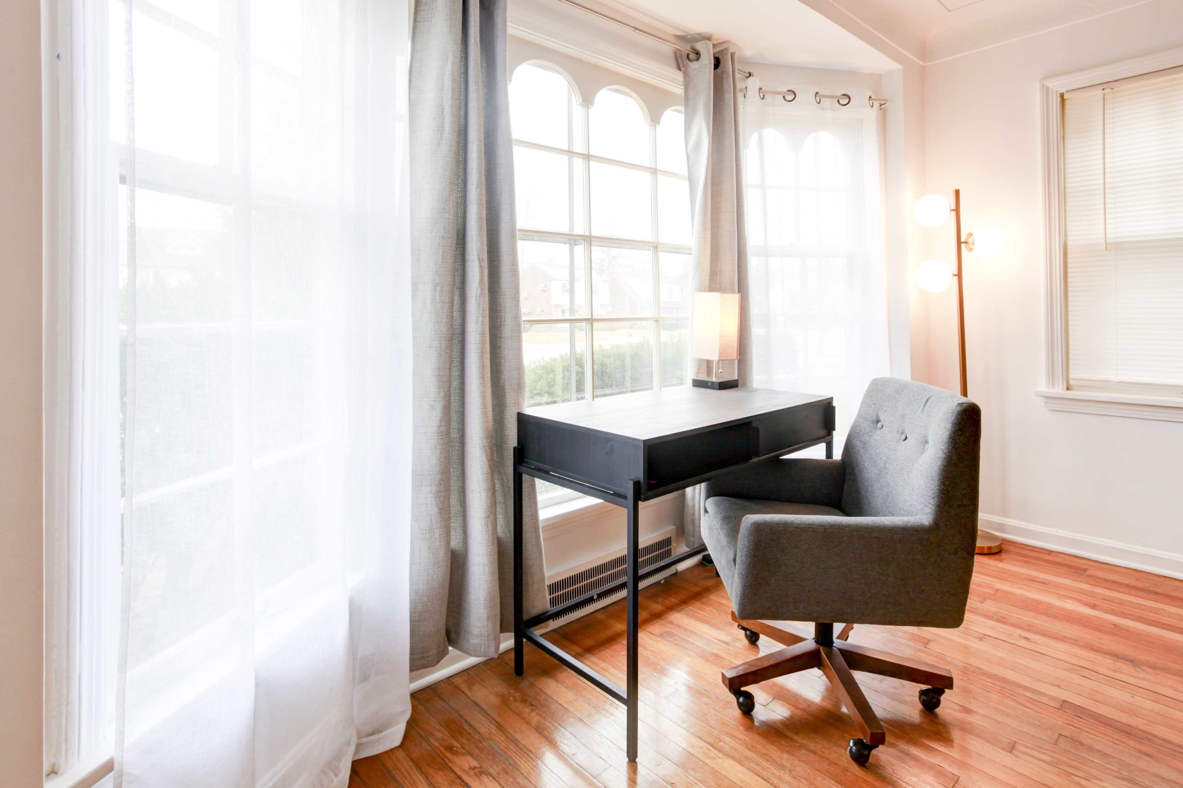 A minimalist office space features a grey chair and a black desk positioned beside large windows, with sheer curtains and warm wood flooring.