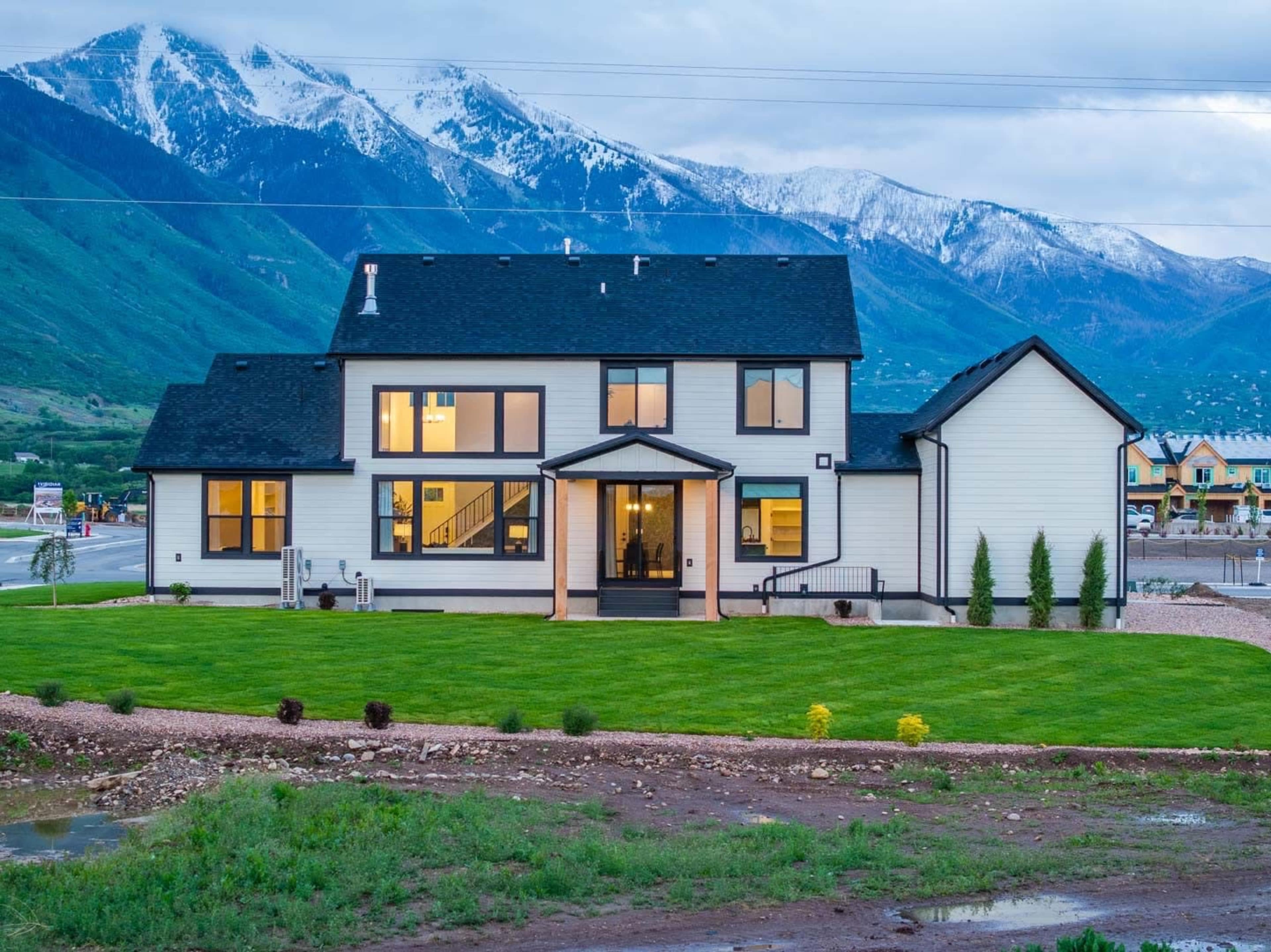 The image shows a modern two-story house with a black roof and large windows, set against a backdrop of snow-capped mountains.