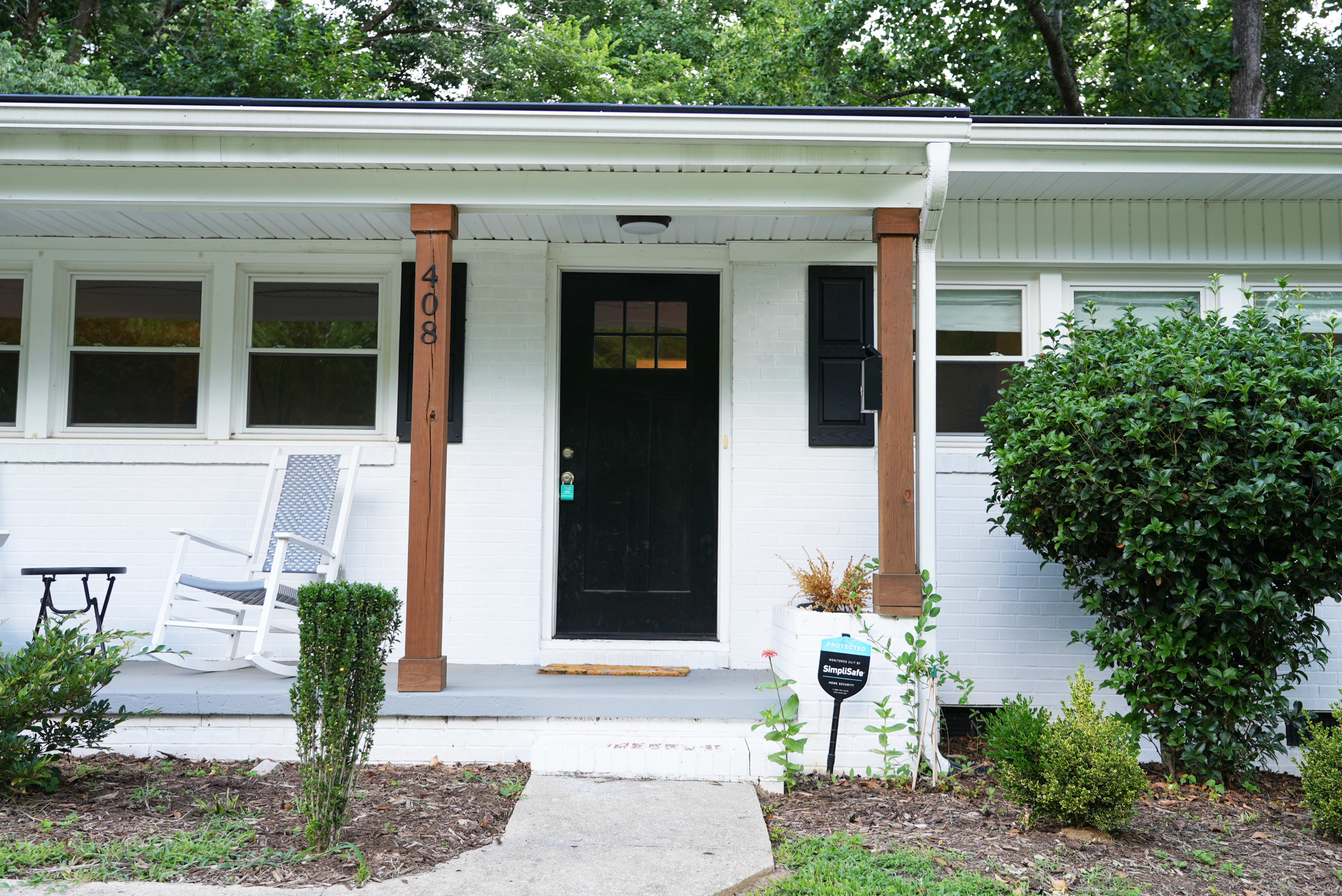 A white house with a black front door, wooden columns, a porch with a rocking chair, and neatly trimmed bushes flanking the entrance.