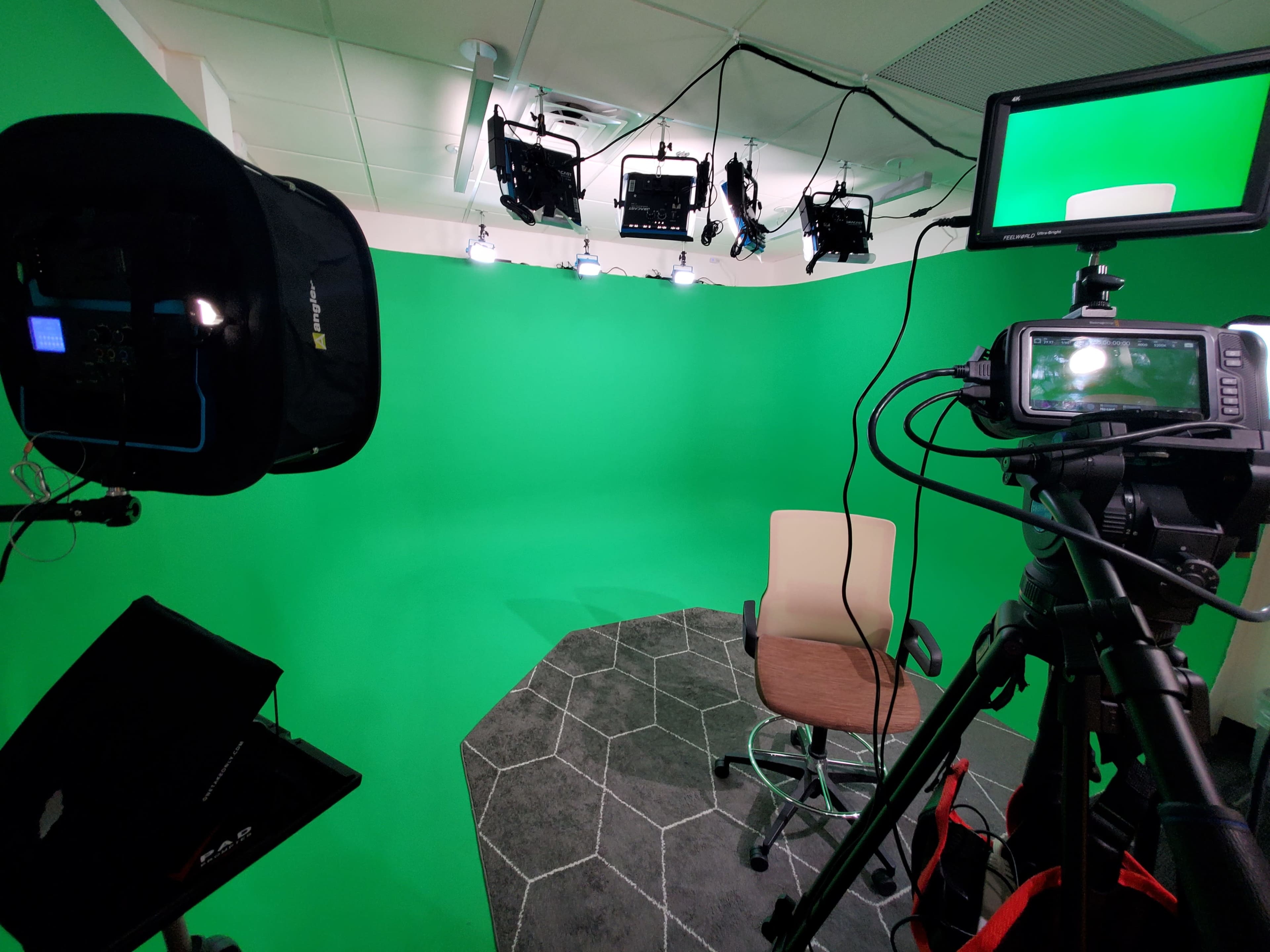The image shows a green screen studio setup with camera equipment and a single chair positioned on a hexagonal-patterned floor.