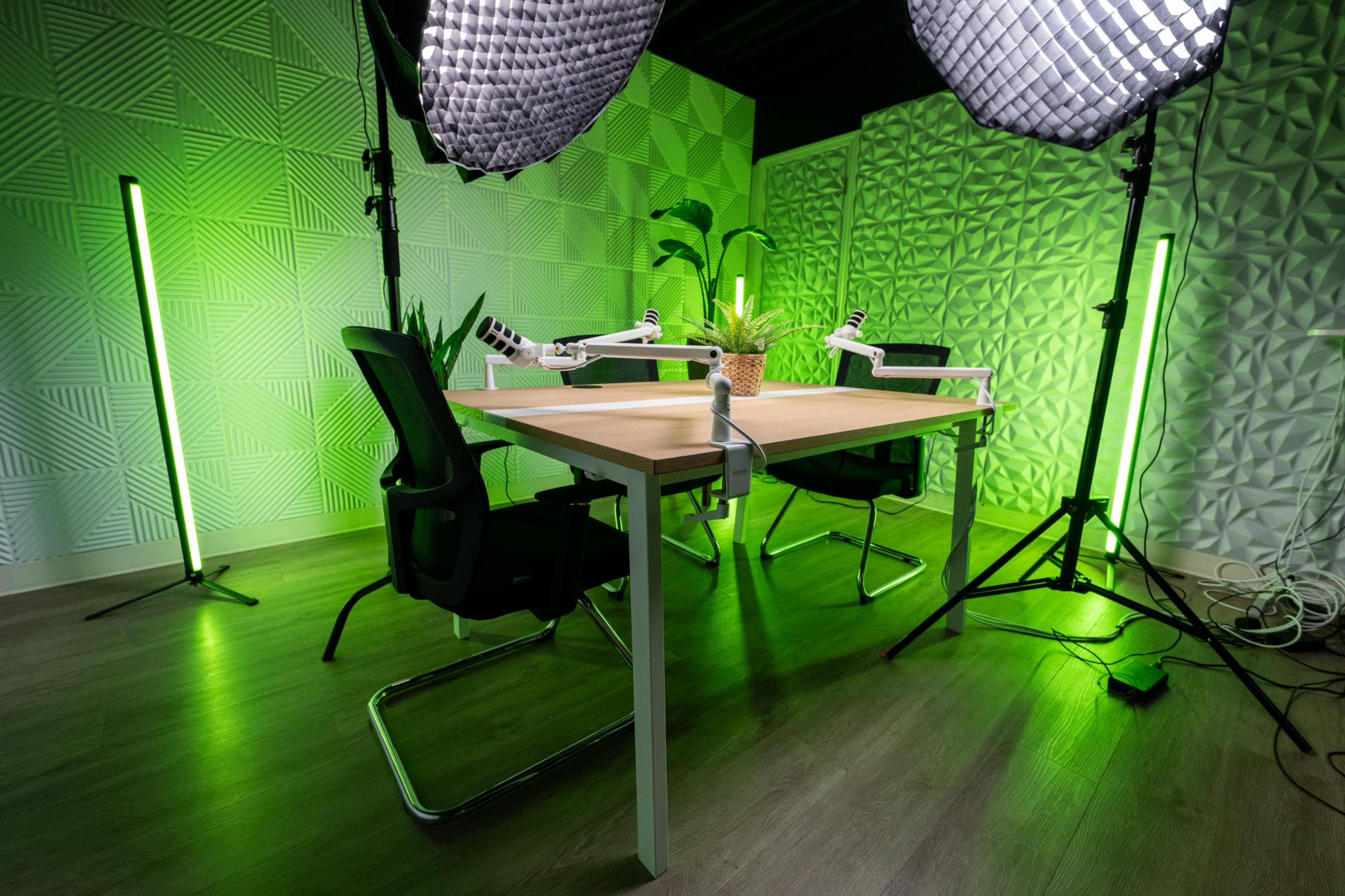 The image shows a modern studio setup featuring a wooden table, two black chairs, and green lighting, with softboxes positioned to illuminate the space and potted plants decoratively arranged.