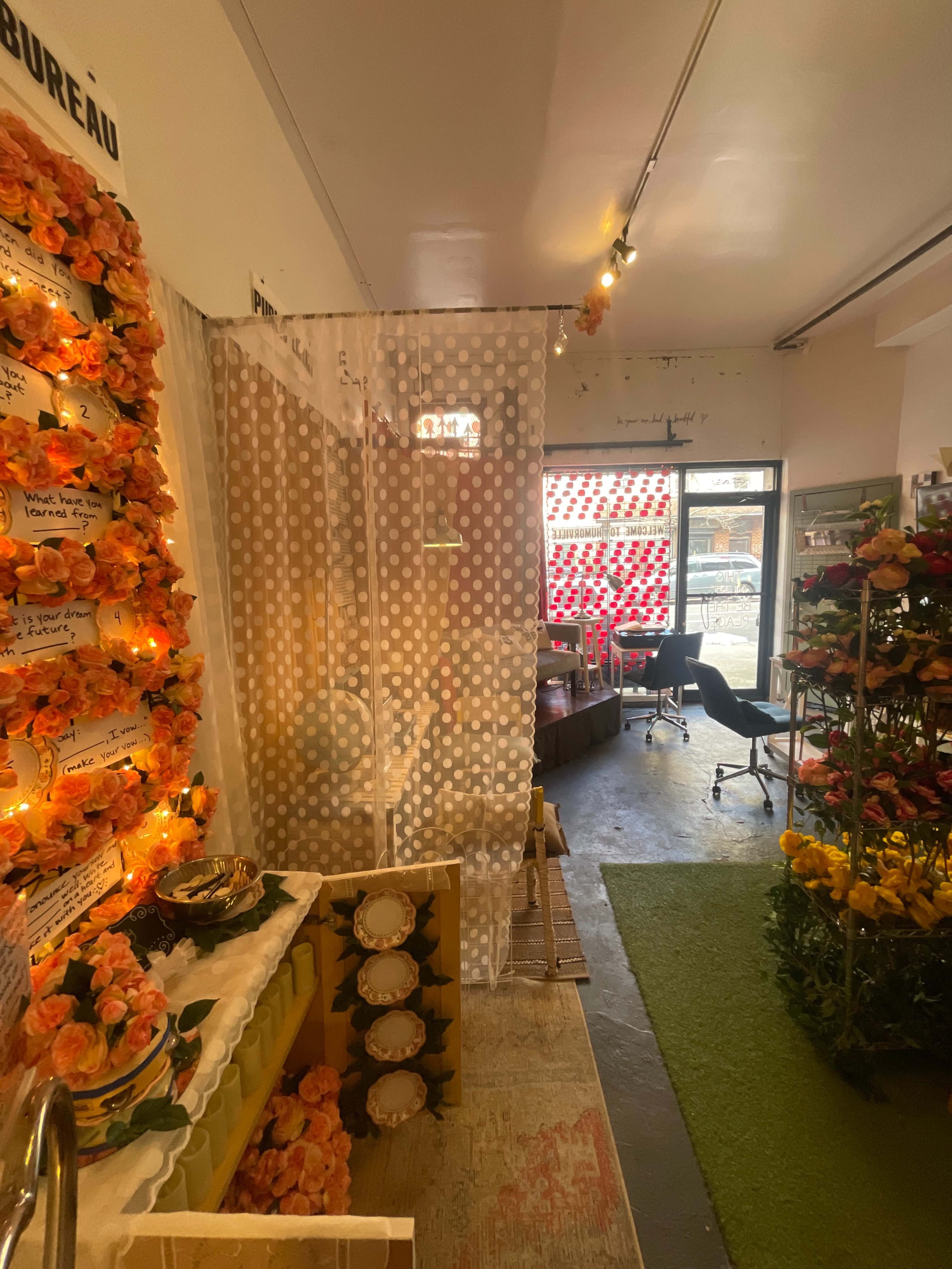 The image shows a brightly decorated interior space featuring flower arrangements, a partition made of sheer fabric, and a glimpse of a workspace with a chair and computer.