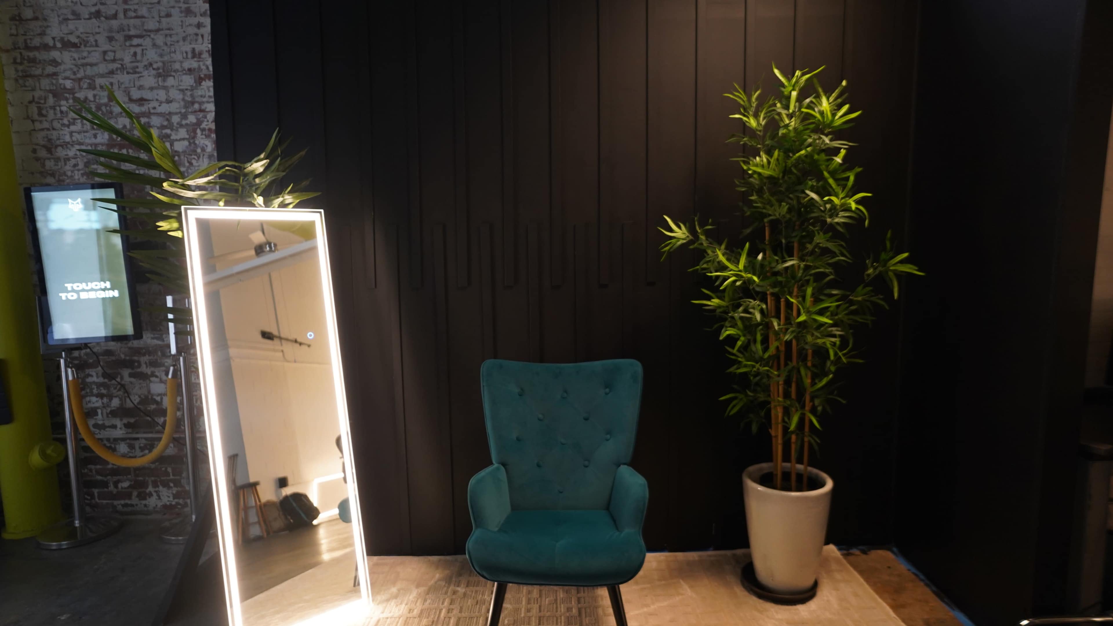 The image features a teal upholstered chair next to a lit mirror and a potted plant against a dark wooden wall.
