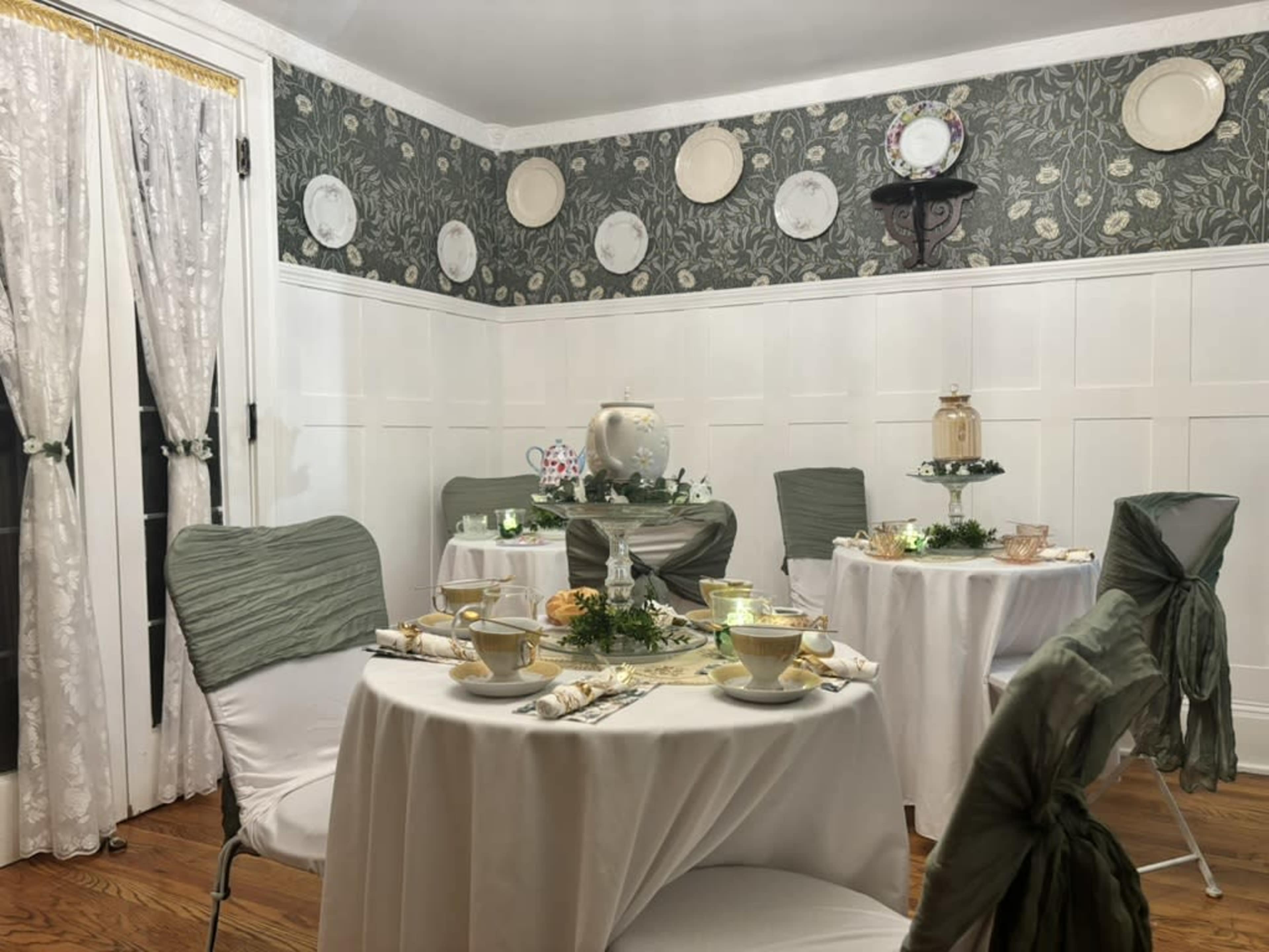 A dining room is set with white tables and chairs adorned with green fabric, and decorative plates hang on the walls.