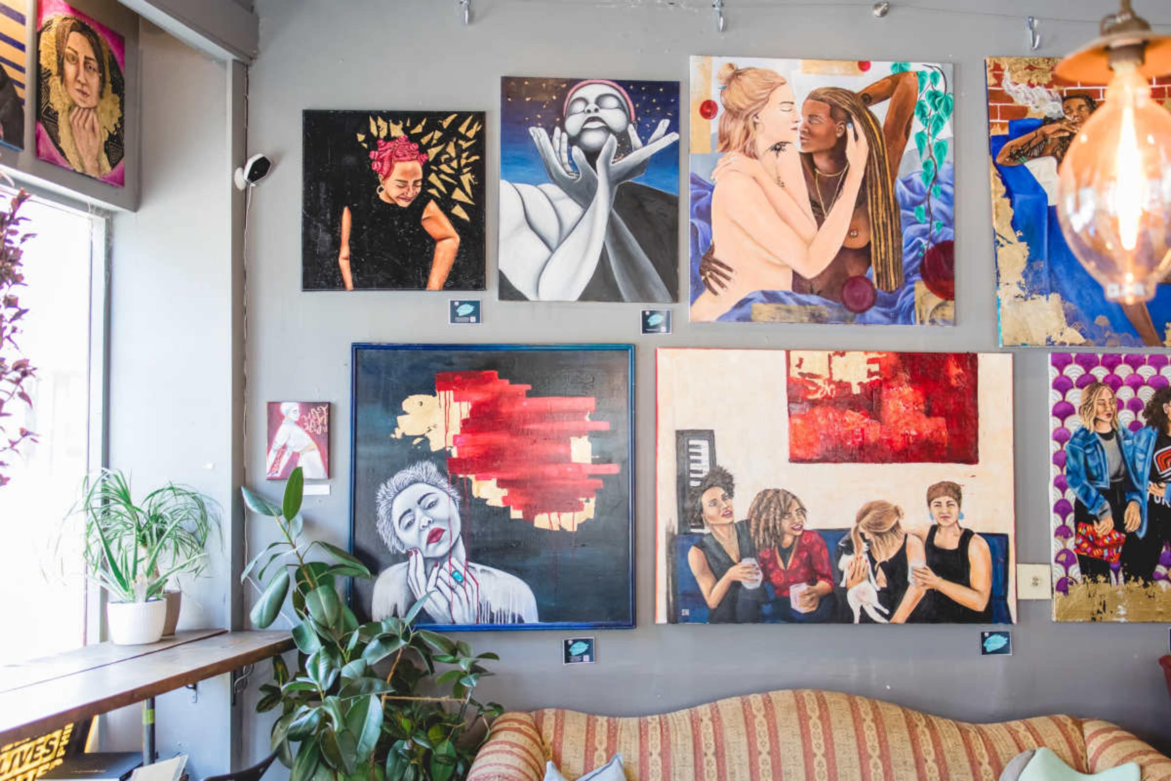 The image shows a cozy indoor space featuring a variety of colorful artworks displayed on the walls, with a sofa and plants in the foreground.