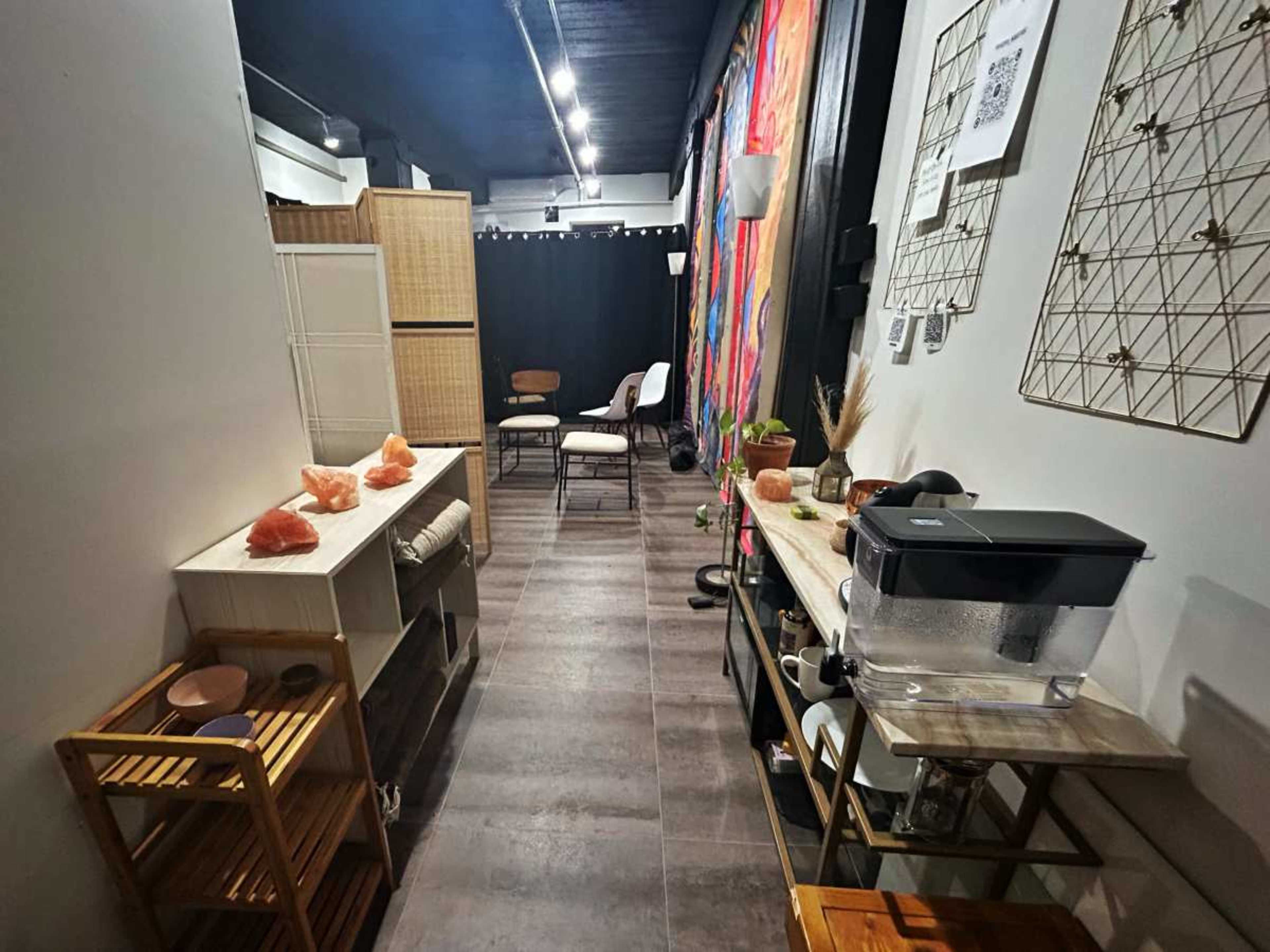 The image shows a narrow, well-decorated room with wooden carts, a water dispenser, and colorful wall hangings, leading to a seating area at the back.