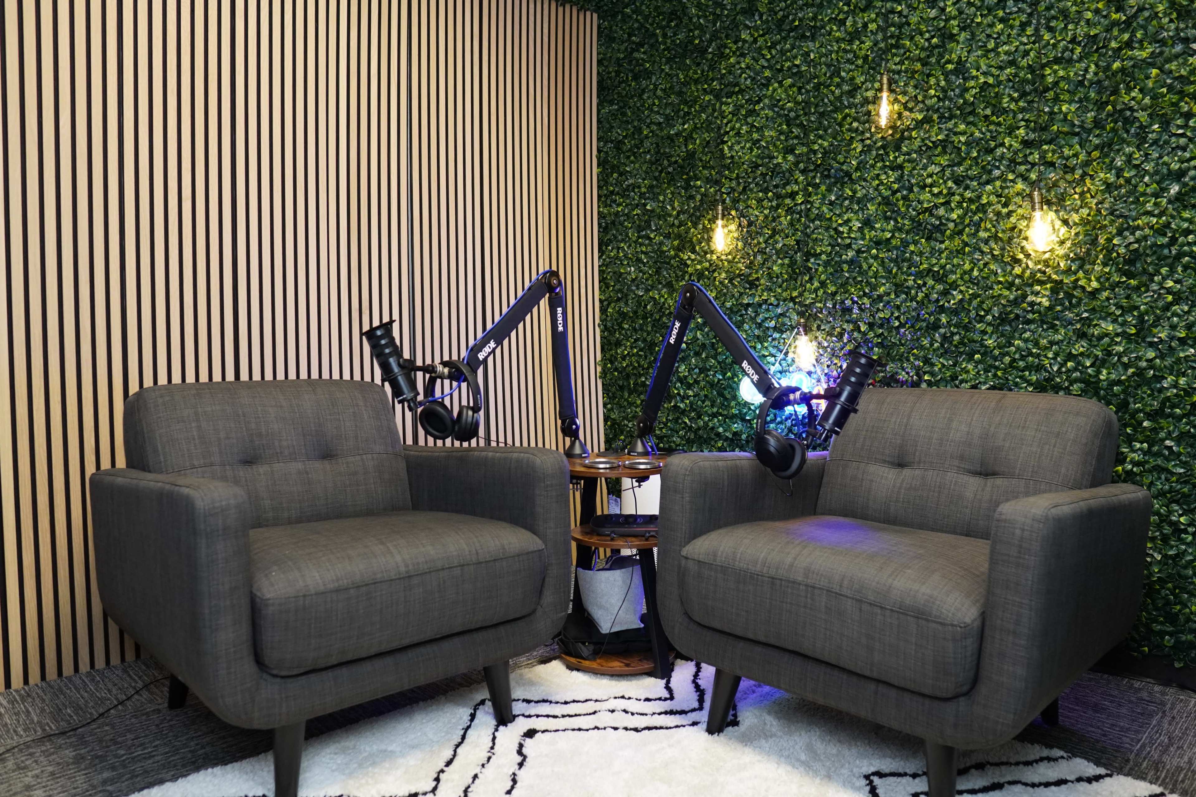 Two gray armchairs are positioned in a small studio with a green wall, equipped with adjustable microphone arms and hanging lights.