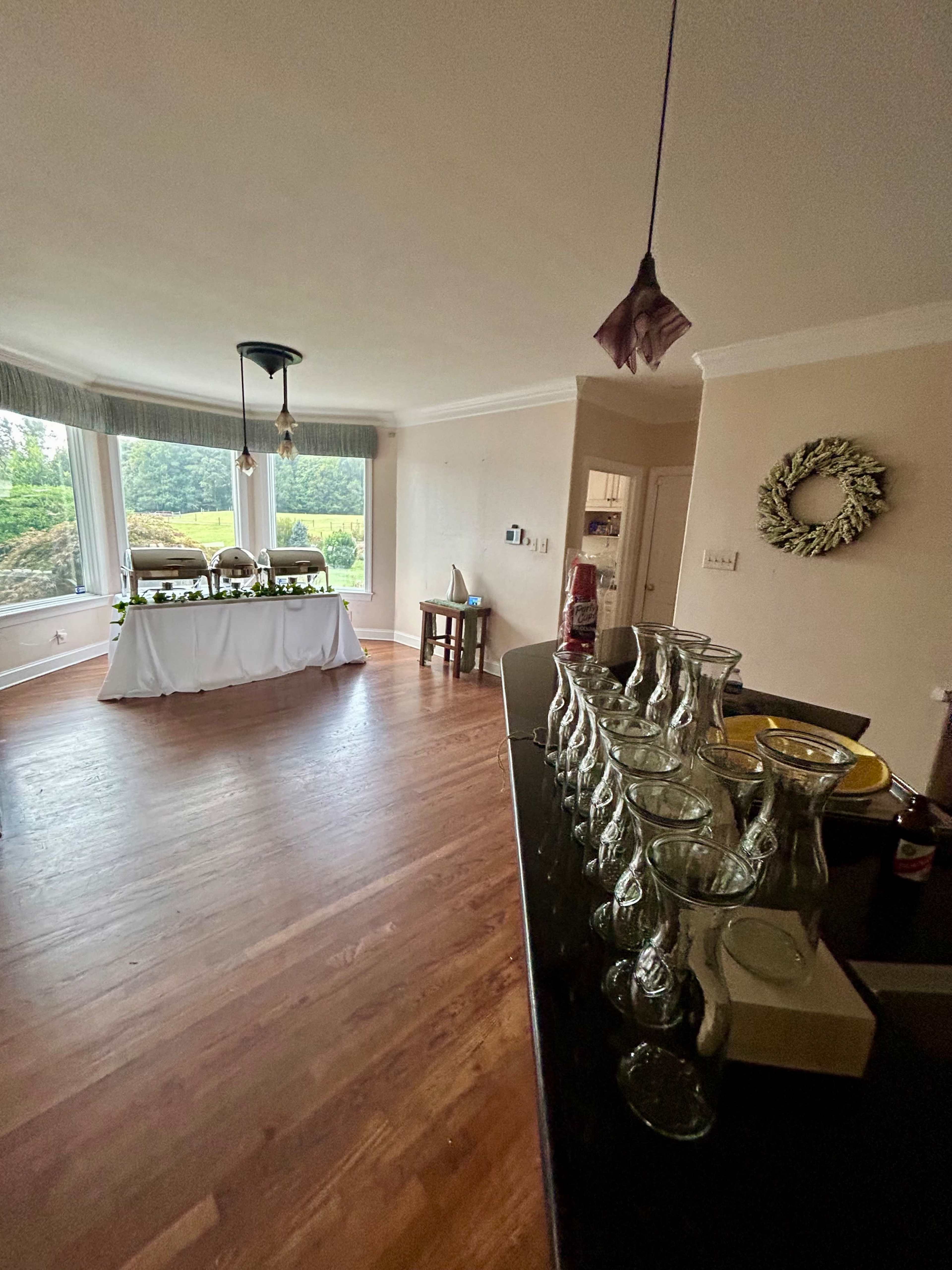 The image shows a spacious, well-lit room with wooden flooring, featuring a table with decorative items and a row of glass bottles lined up on a counter.