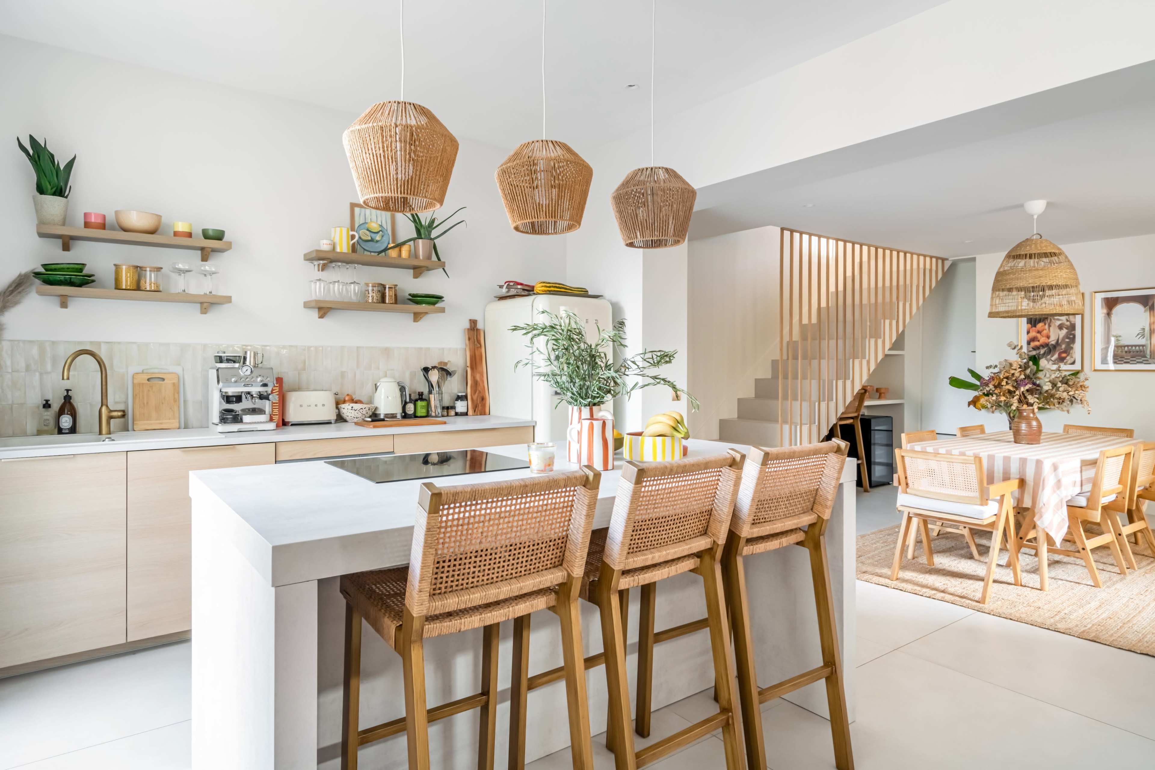 A modern kitchen features light wood cabinetry, woven pendant lights, and a dining space with a table set for six.