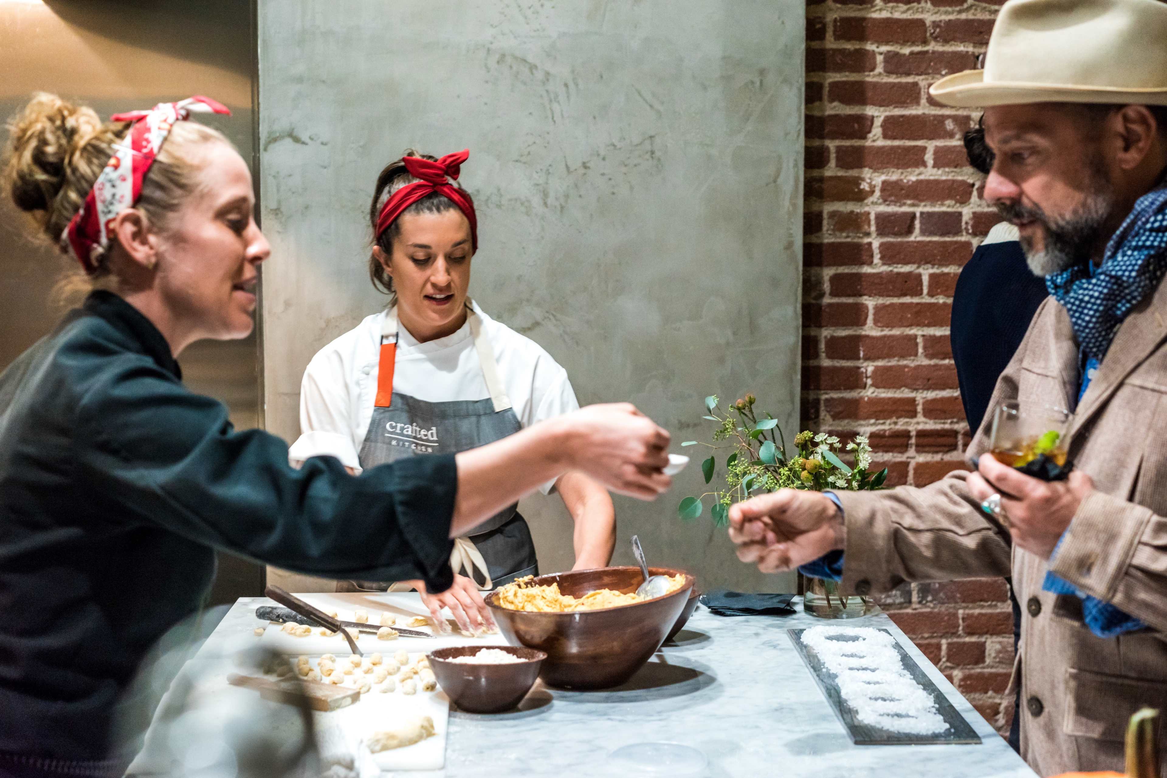 Three people are interacting in a kitchen, with two women preparing food and a man observing while holding a small dish.