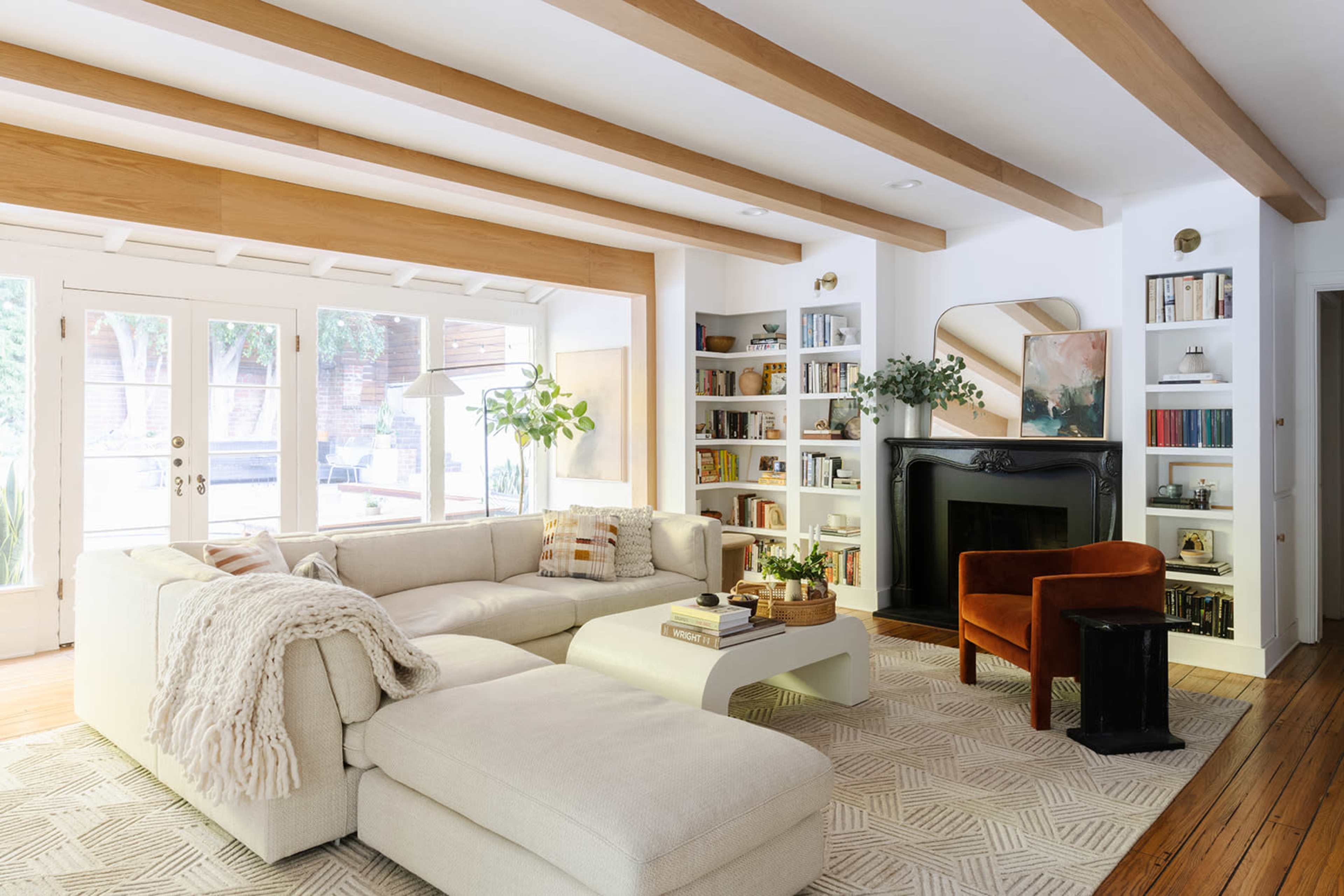 The living room features a large white sectional sofa, a coffee table, a cozy chair, bookshelves, and a fireplace with a mirror above it, all set against a backdrop of wooden beams and natural light from the windows.