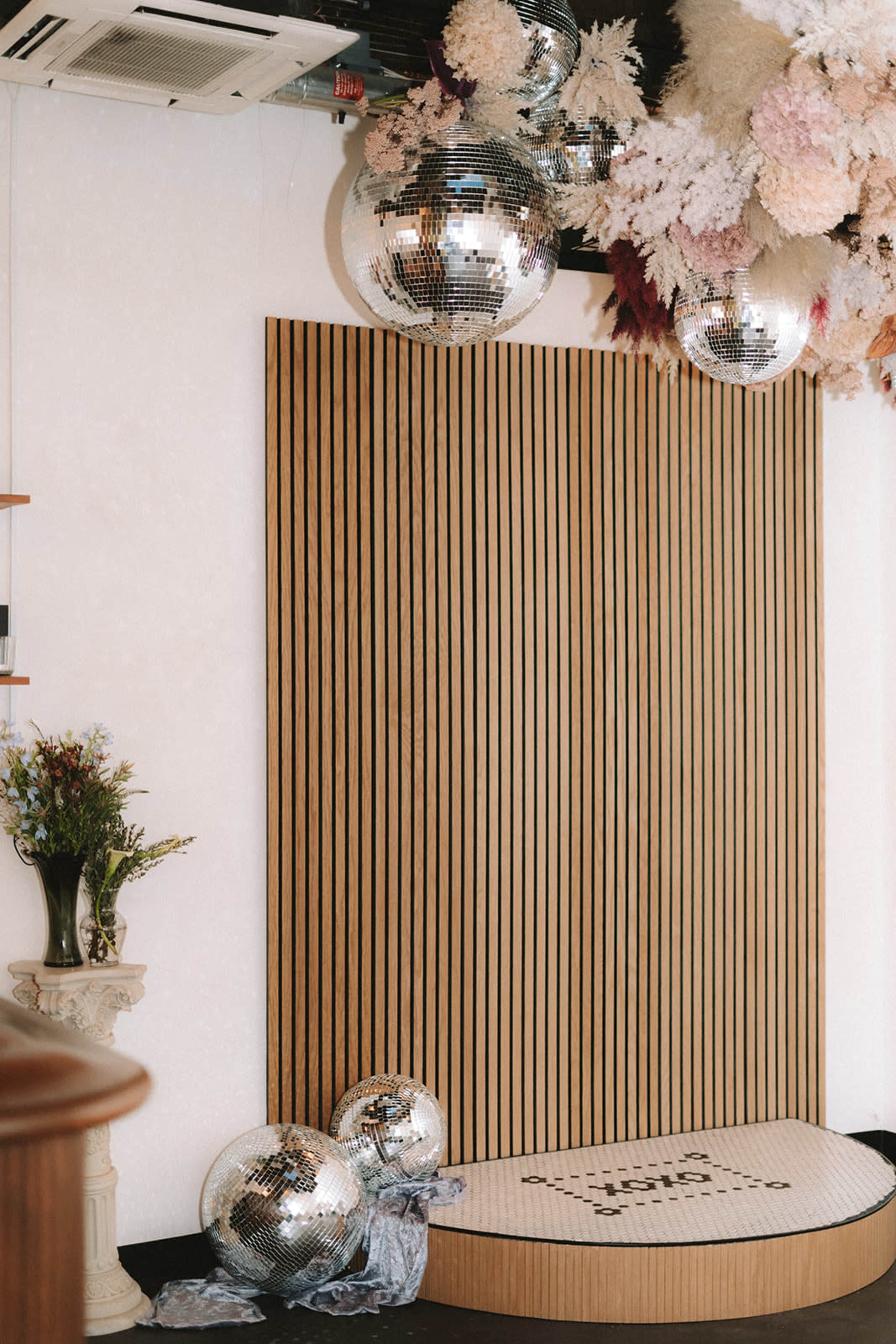 The image features a room with a wooden slatted wall, decorative disco balls hanging from the ceiling, and a stylized floor area with floral arrangements.