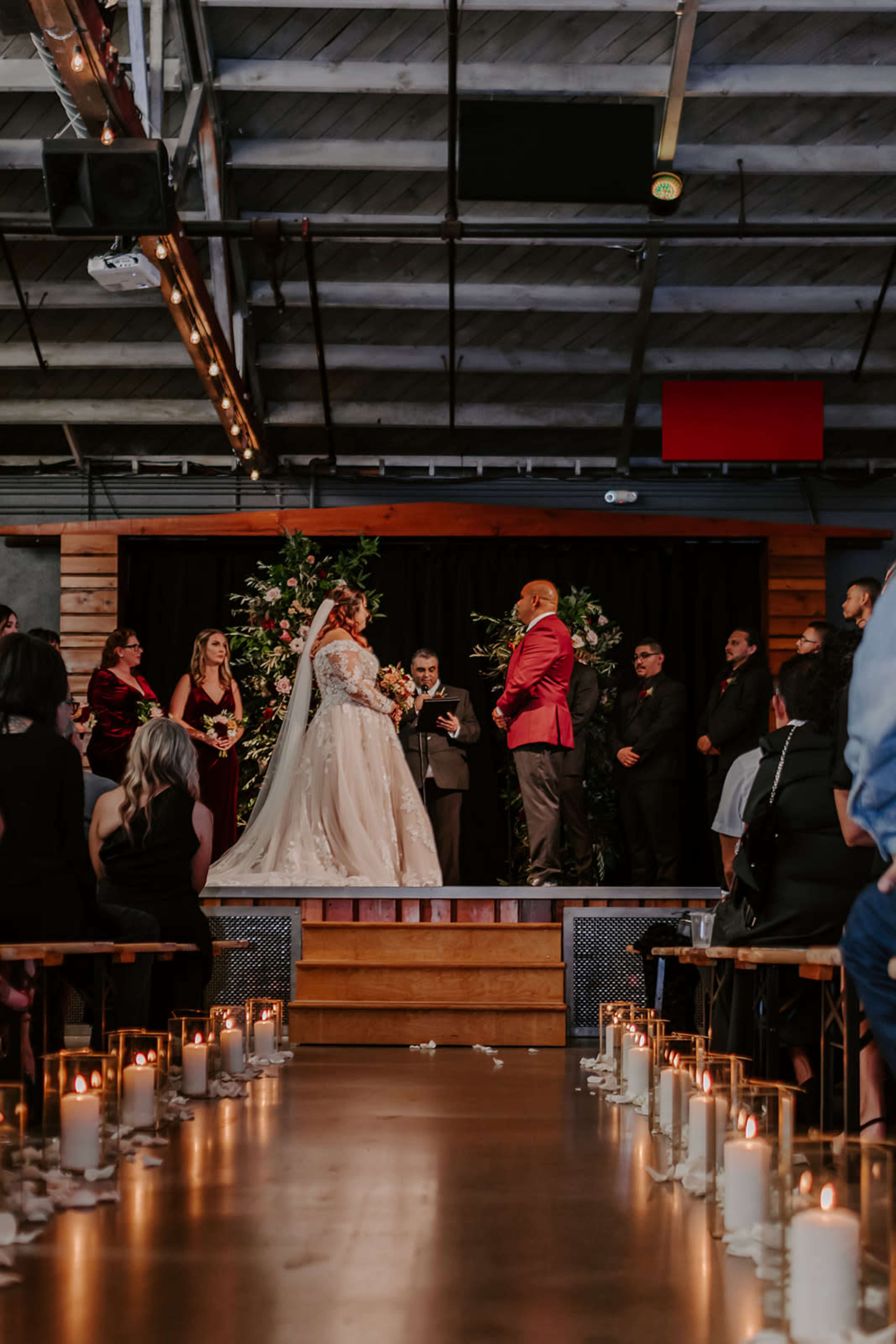 A bride and groom stand at the altar during their wedding ceremony, surrounded by their wedding party and decorated with floral arrangements and candles lining the aisle.