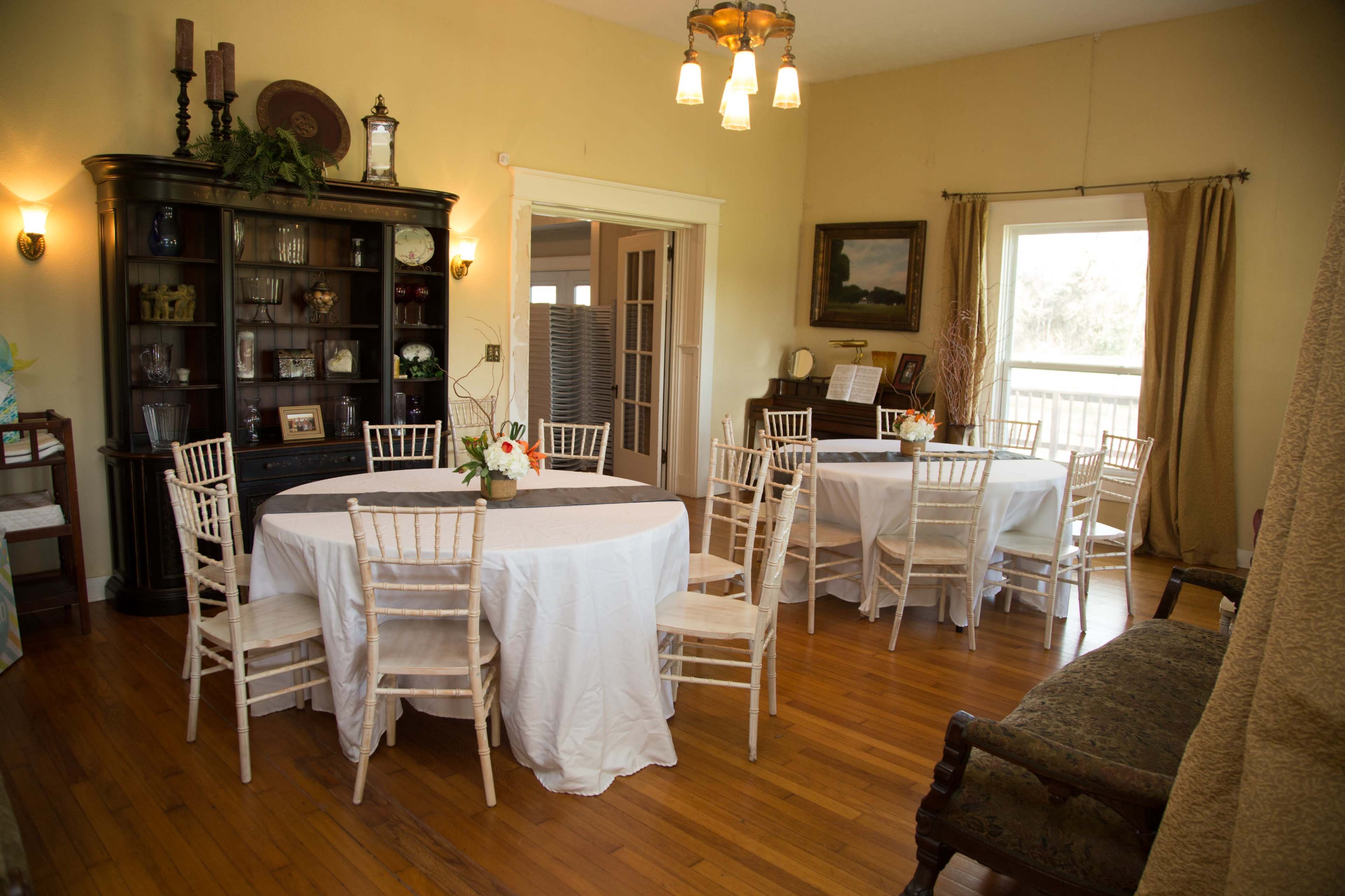 The image shows a spacious dining area with two round tables covered in white tablecloths, surrounded by wooden chairs, and featuring a floral centerpiece.