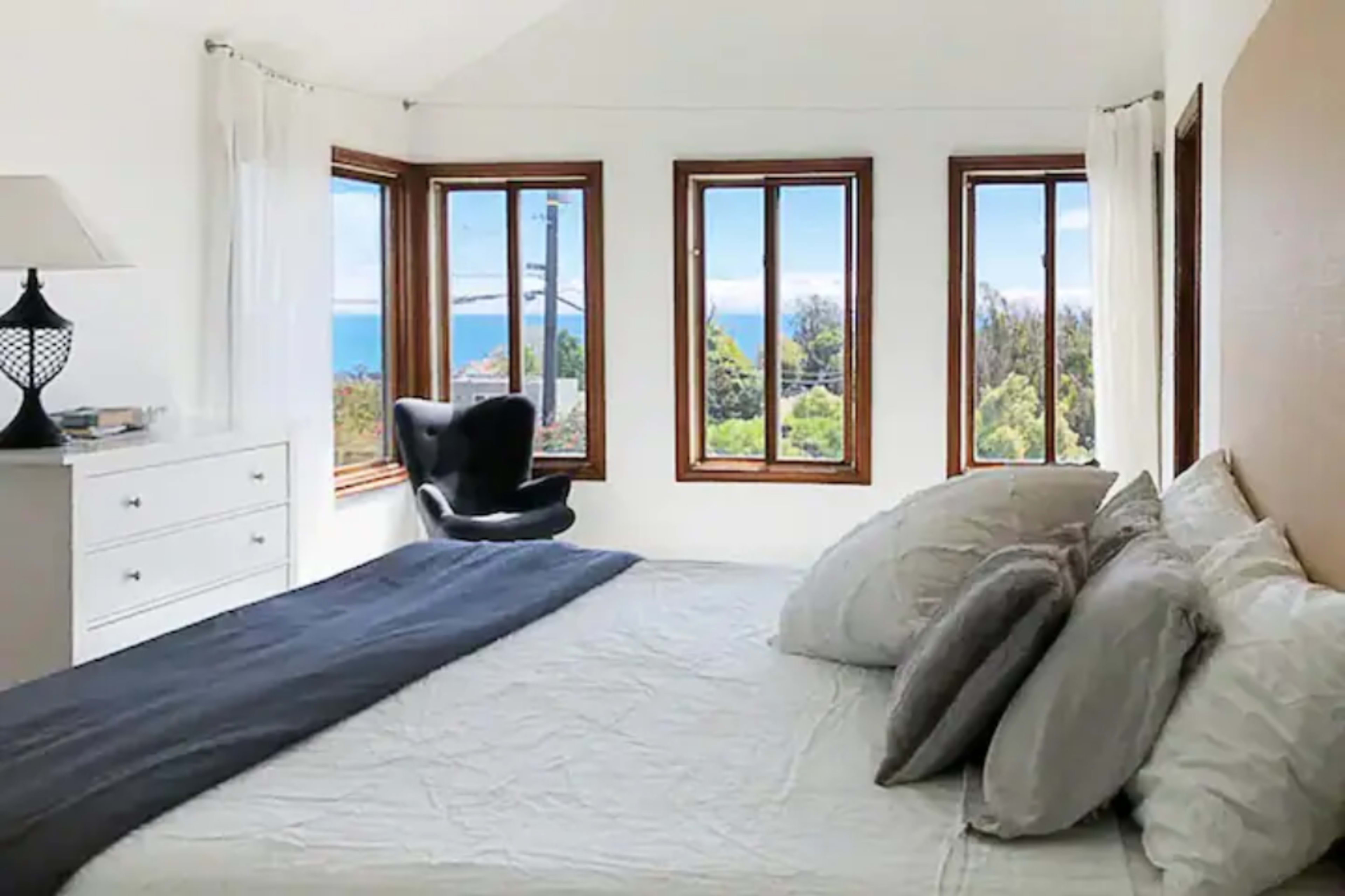 A bright bedroom features a large bed, a black chair by a window, and multiple panes of glass offering a view of the ocean and surrounding greenery.