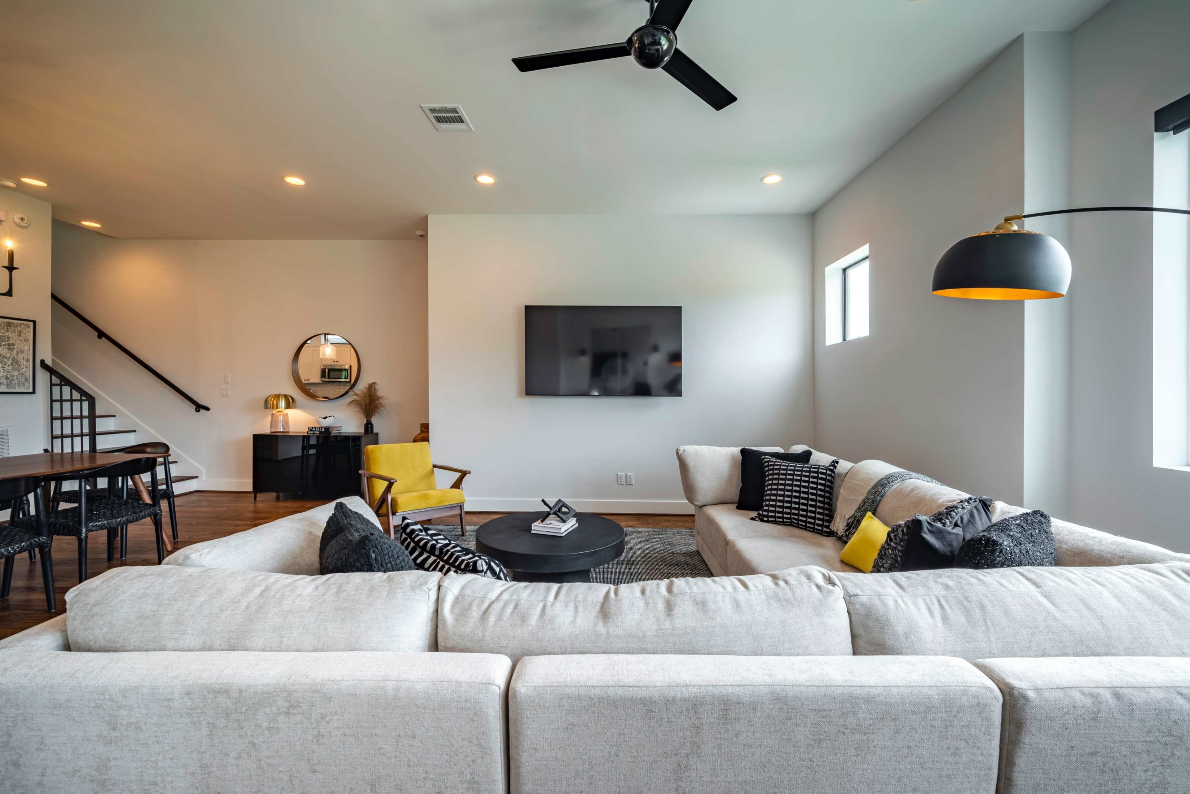 A modern living room features a large sectional sofa, a coffee table, and a mounted television, with a dining area visible in the background.