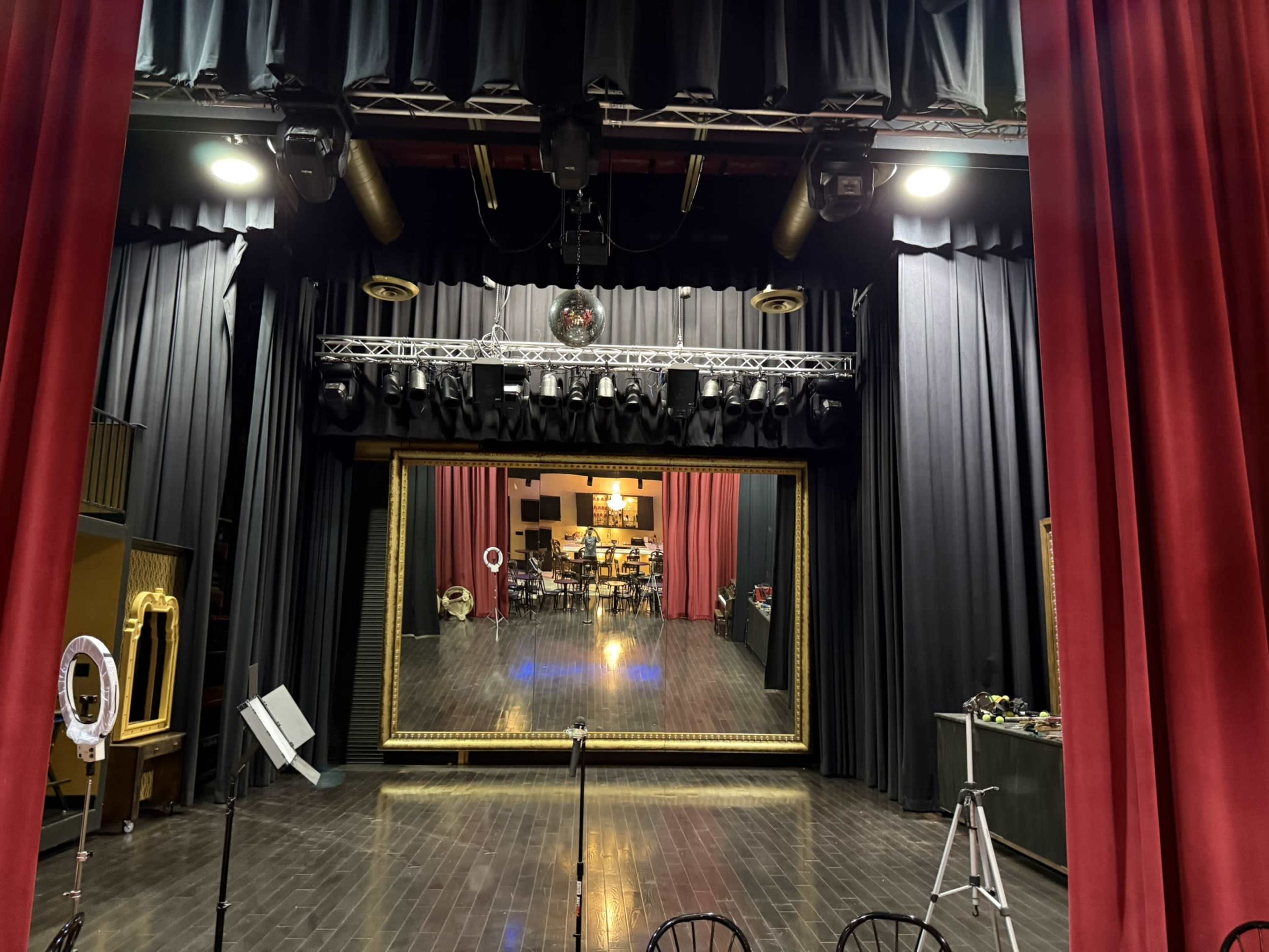 The image shows the interior of a stage setting with red curtains, lights, a large mirror backdrop, and a dimly lit audience area.