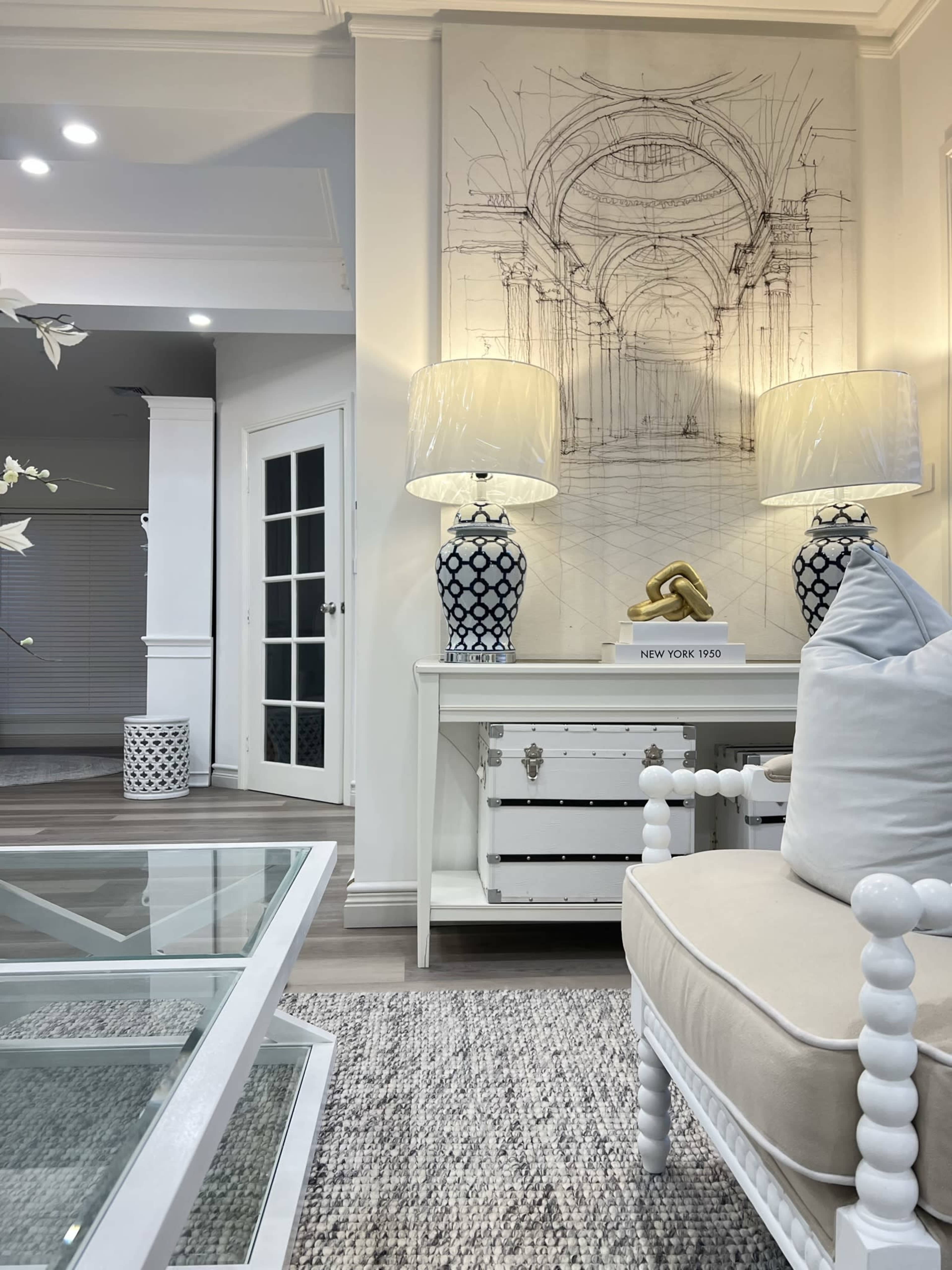A stylish living room featuring a white sofa with light cushions, a glass coffee table, floor lamps with patterned bases, and a decorative wall art piece depicting an architectural sketch.
