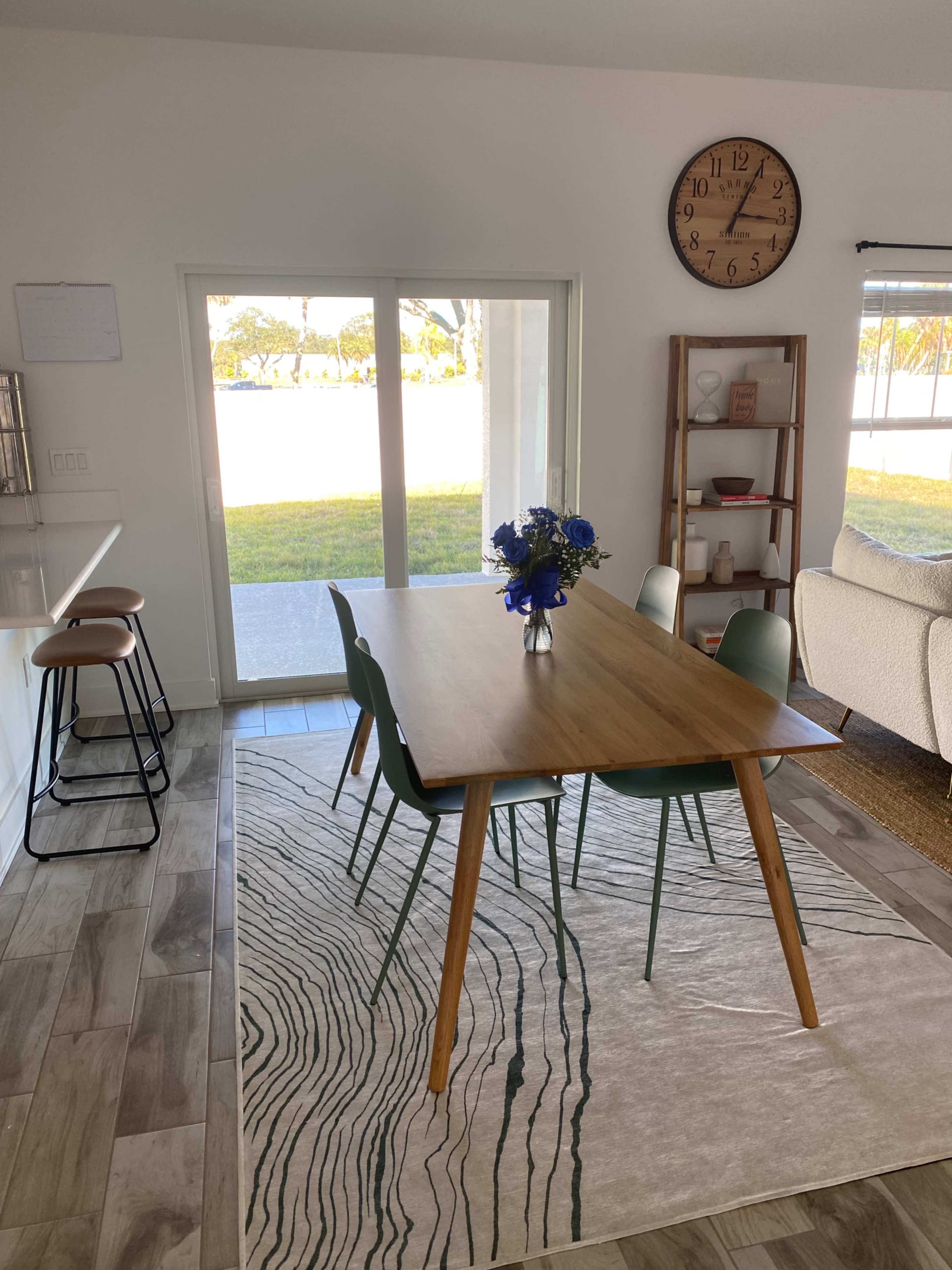 A dining table with four chairs and a vase of flowers sits on a rug in a bright room with large sliding doors leading to a grassy outdoor area.