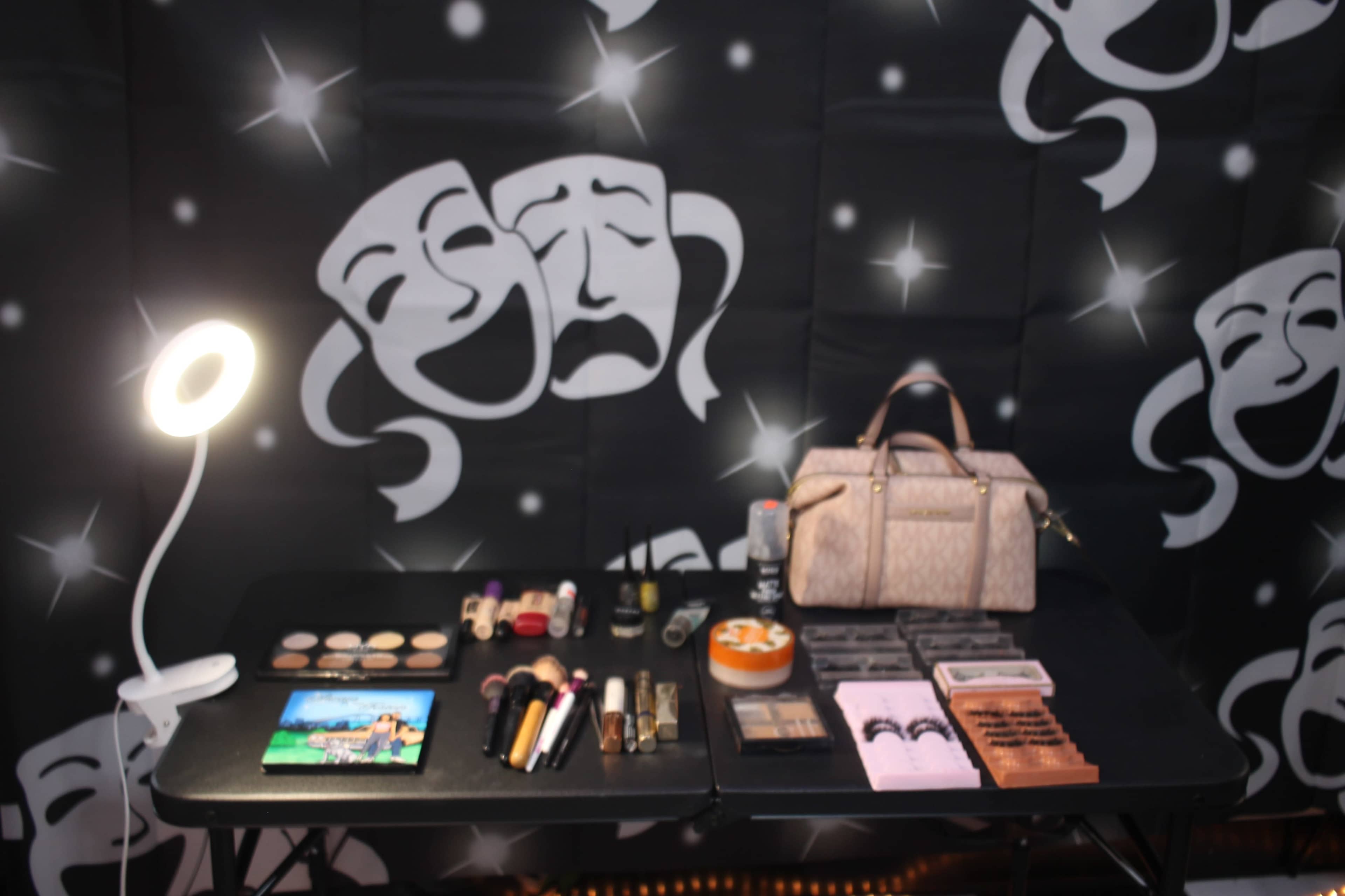 A table displays various makeup products and a handbag against a black backdrop featuring theatrical masks.