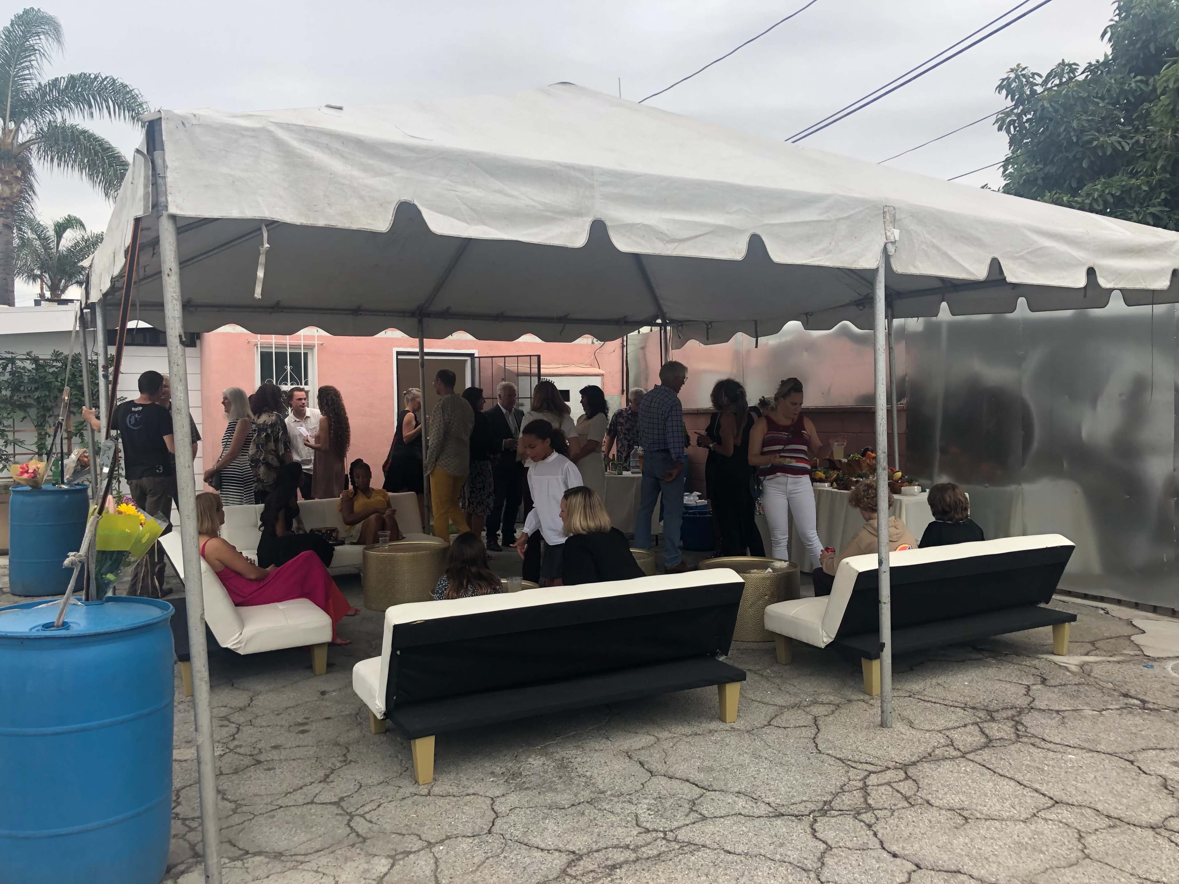A large tent shelters a gathering of people socializing and seated on white sofas in an outdoor space.