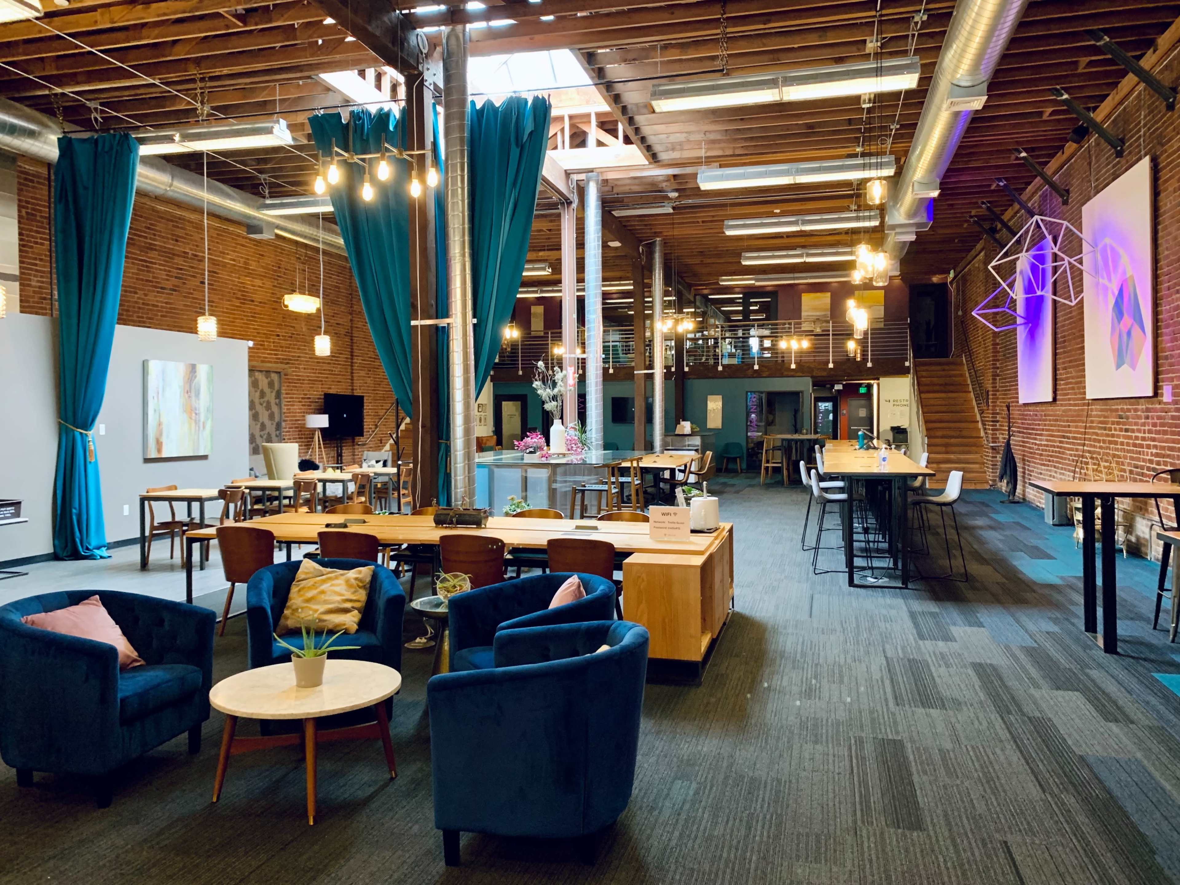 An open, modern interior space with wooden beams, blue curtains, and several seating areas, including couches and tables, illuminated by pendant lights.