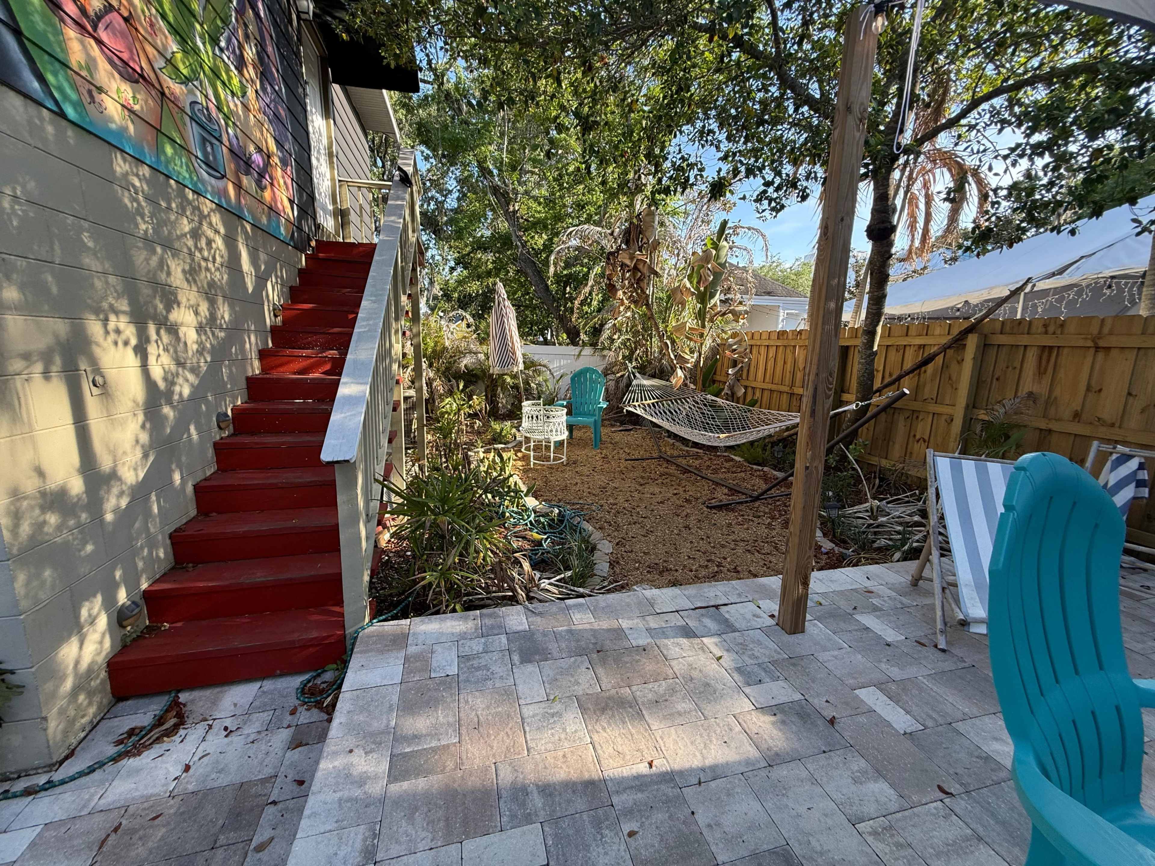 The image shows a patio area with red stairs leading up to a house, surrounded by greenery and featuring a hammock and two chairs on a sandy ground.