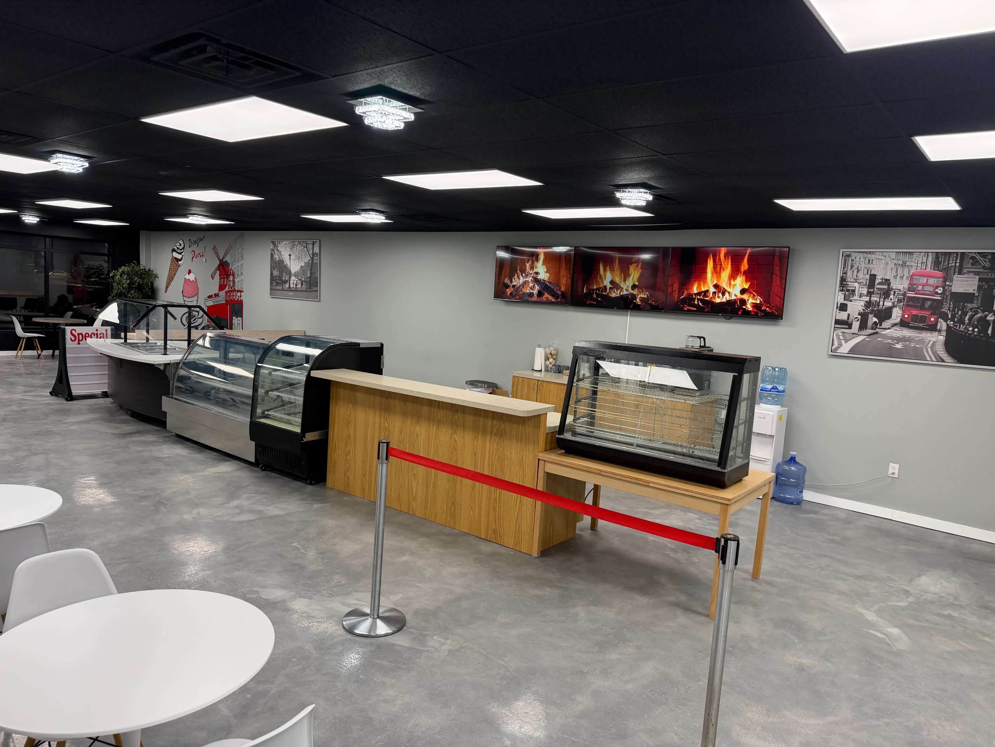 The image shows a modern café interior featuring a service counter with glass display cases, a wooden section, and a seating area with round tables.