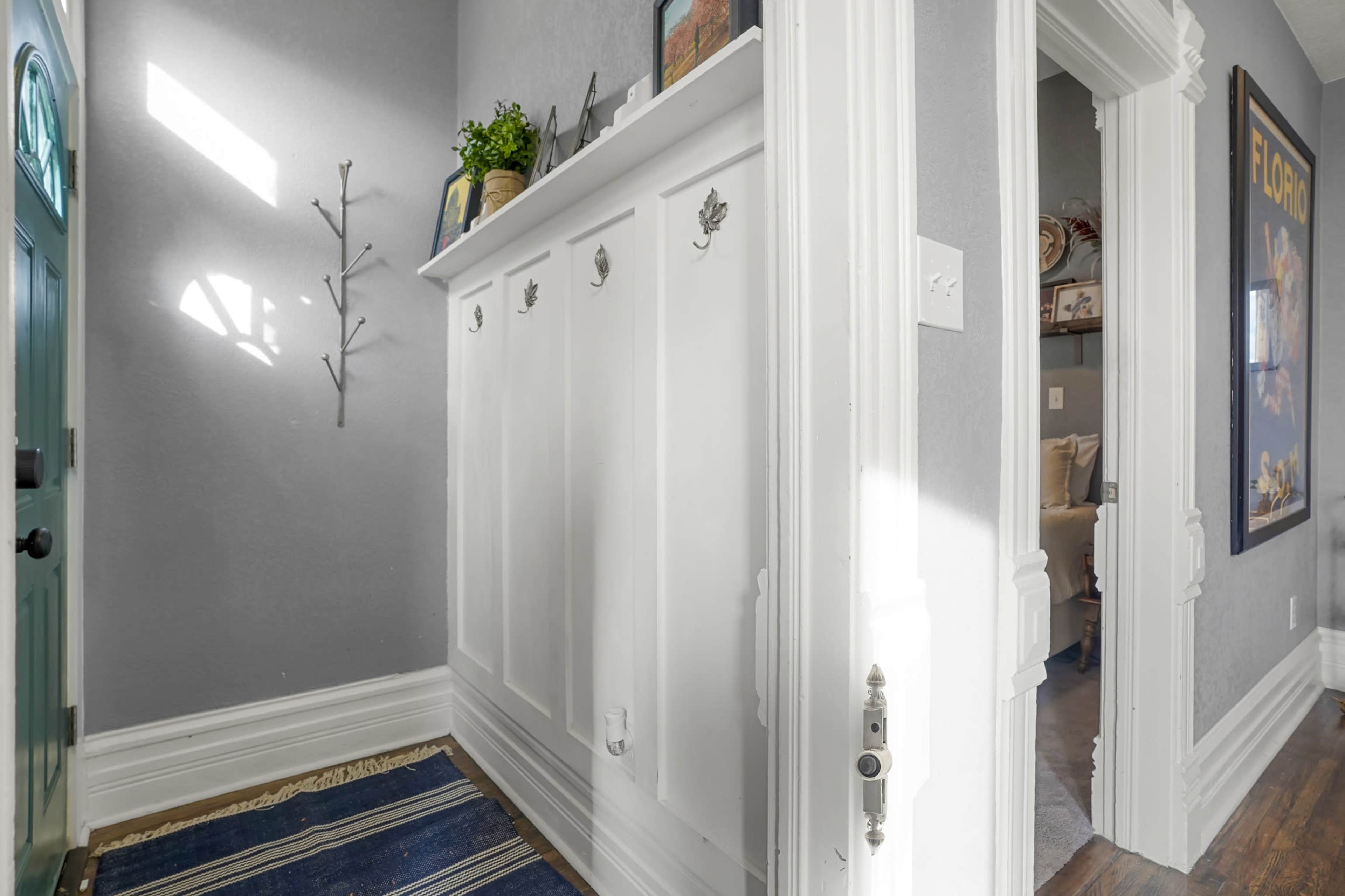 A bright hallway featuring a white wall cabinet with hooks, a door leading to another room, and light streaming through a window.