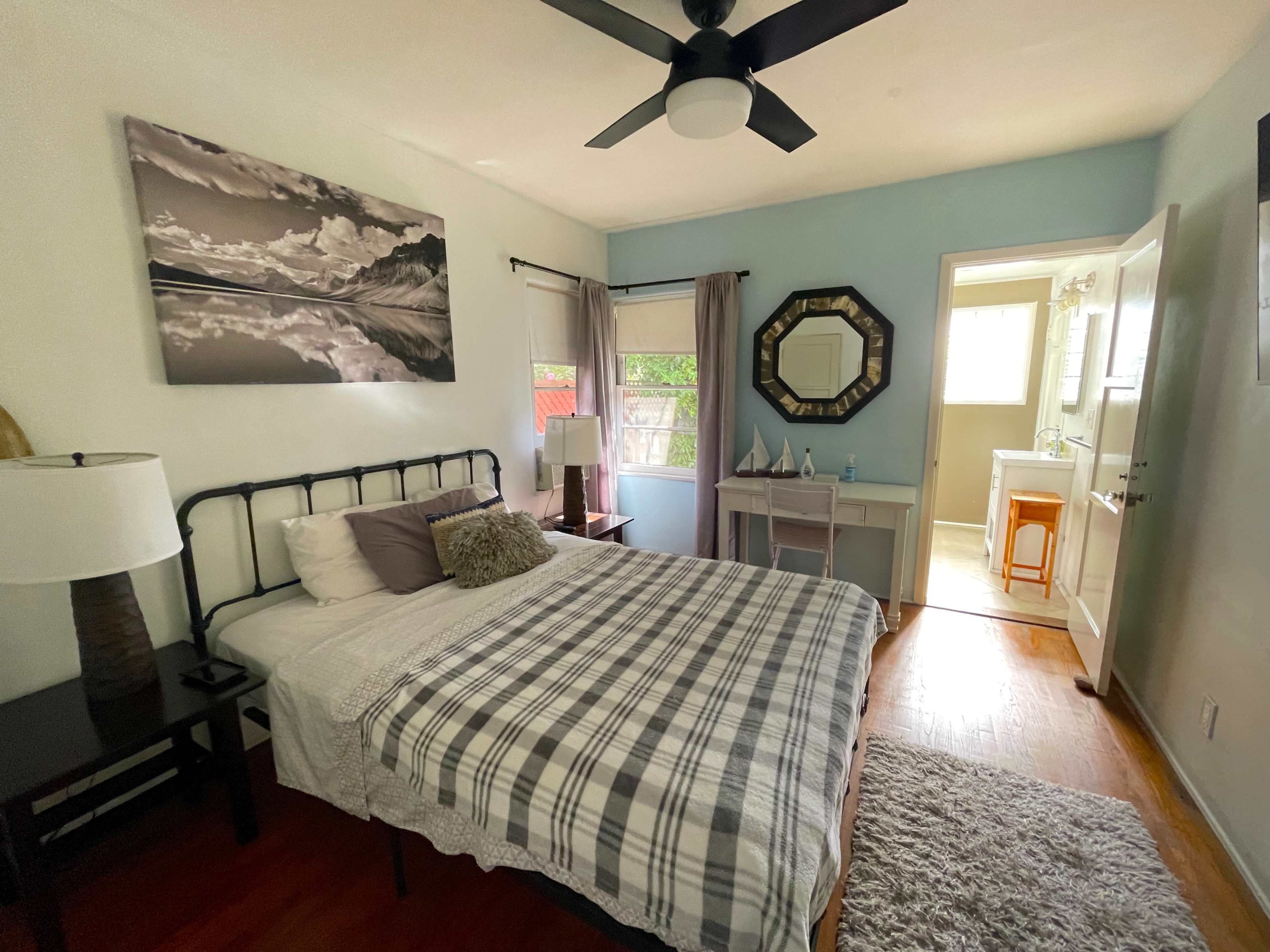 The image shows a well-lit bedroom featuring a bed with a checkered blanket, a nightstand with a lamp, a vanity area, and an en-suite bathroom visible in the background.