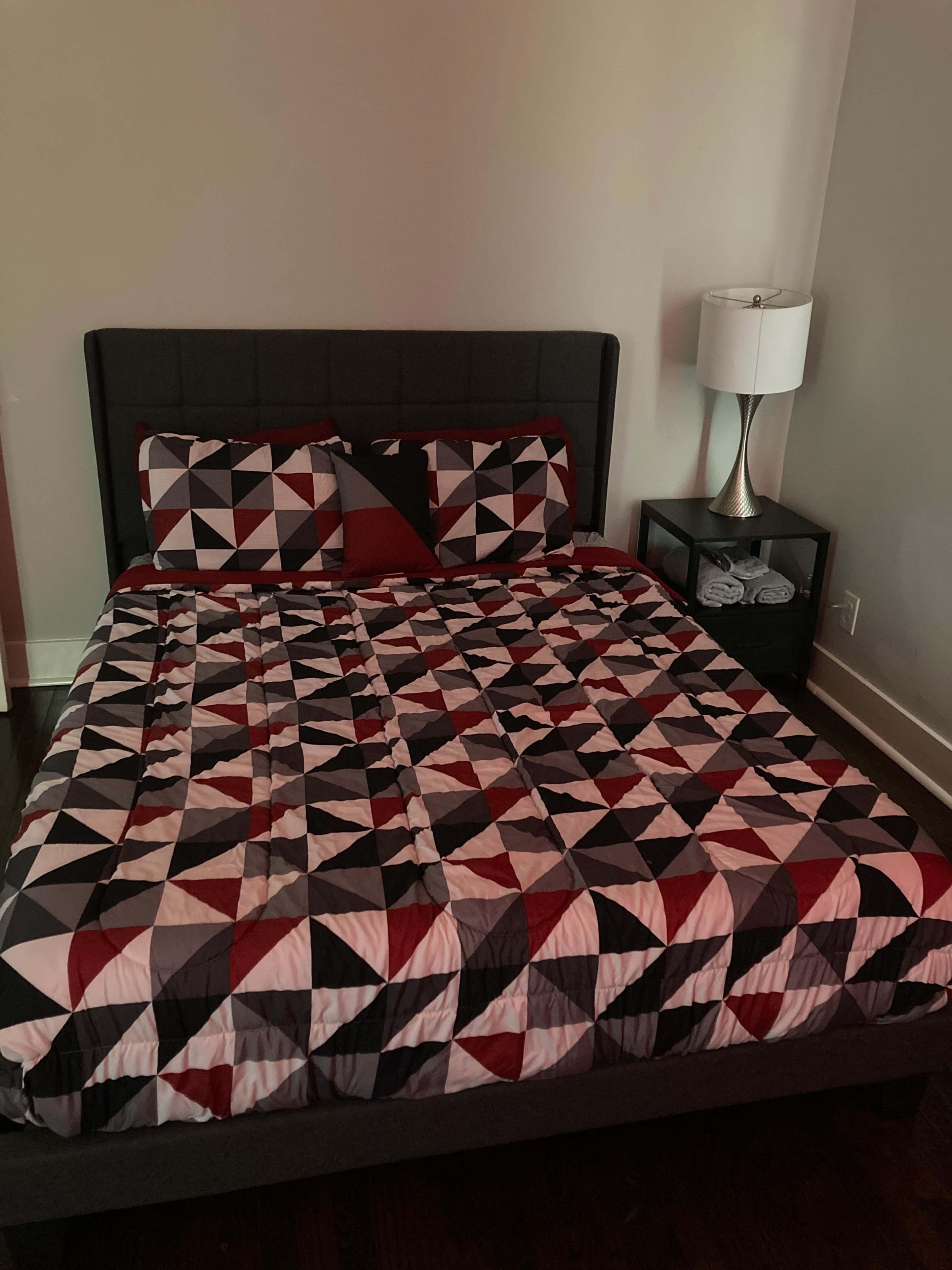 The image shows a neatly made bed with a geometric patterned quilt in red, black, and white, positioned next to a nightstand containing folded towels and a lamp.