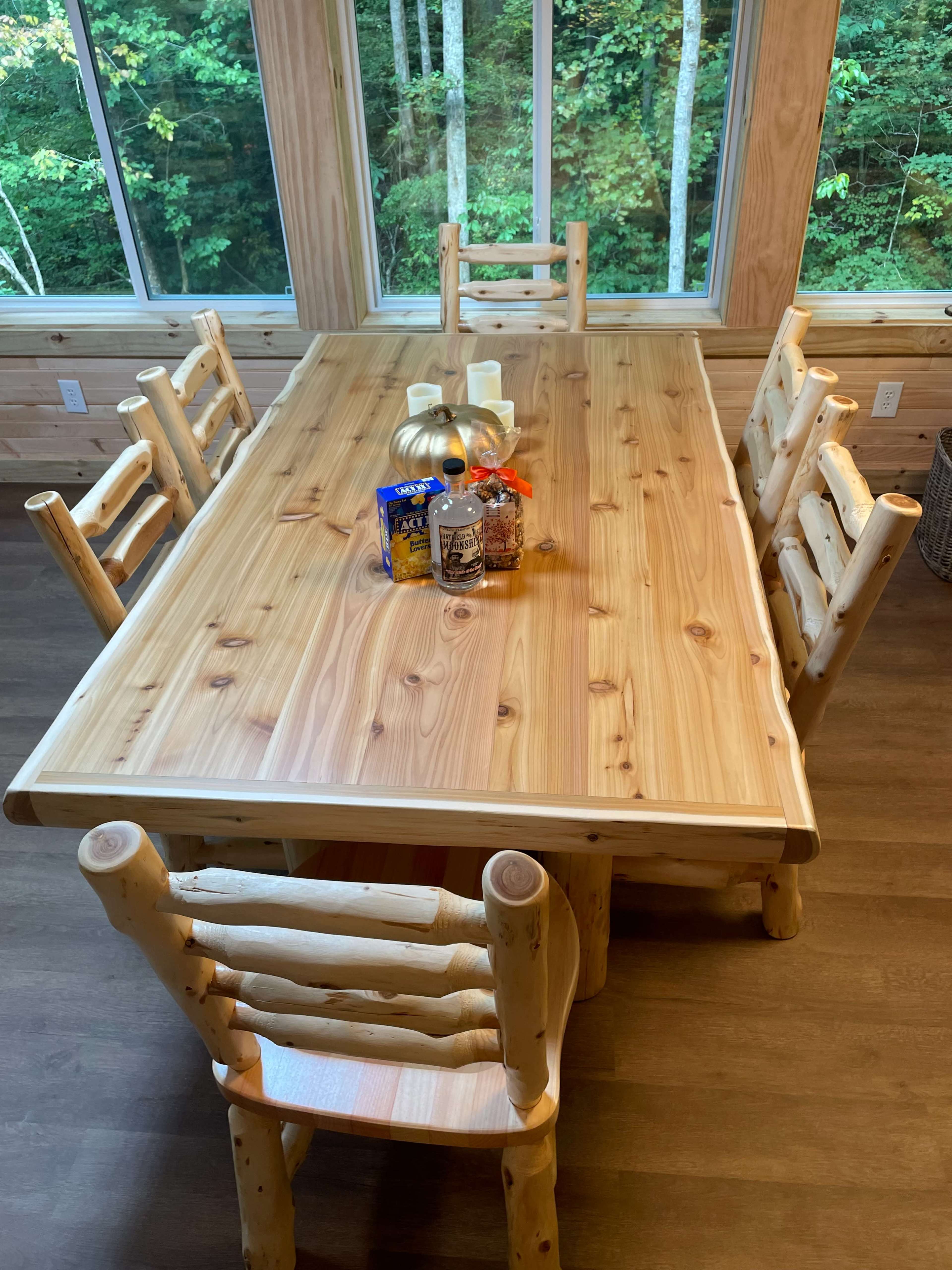 A large wooden dining table surrounded by six matching chairs is set in a room with windows overlooking a wooded area.