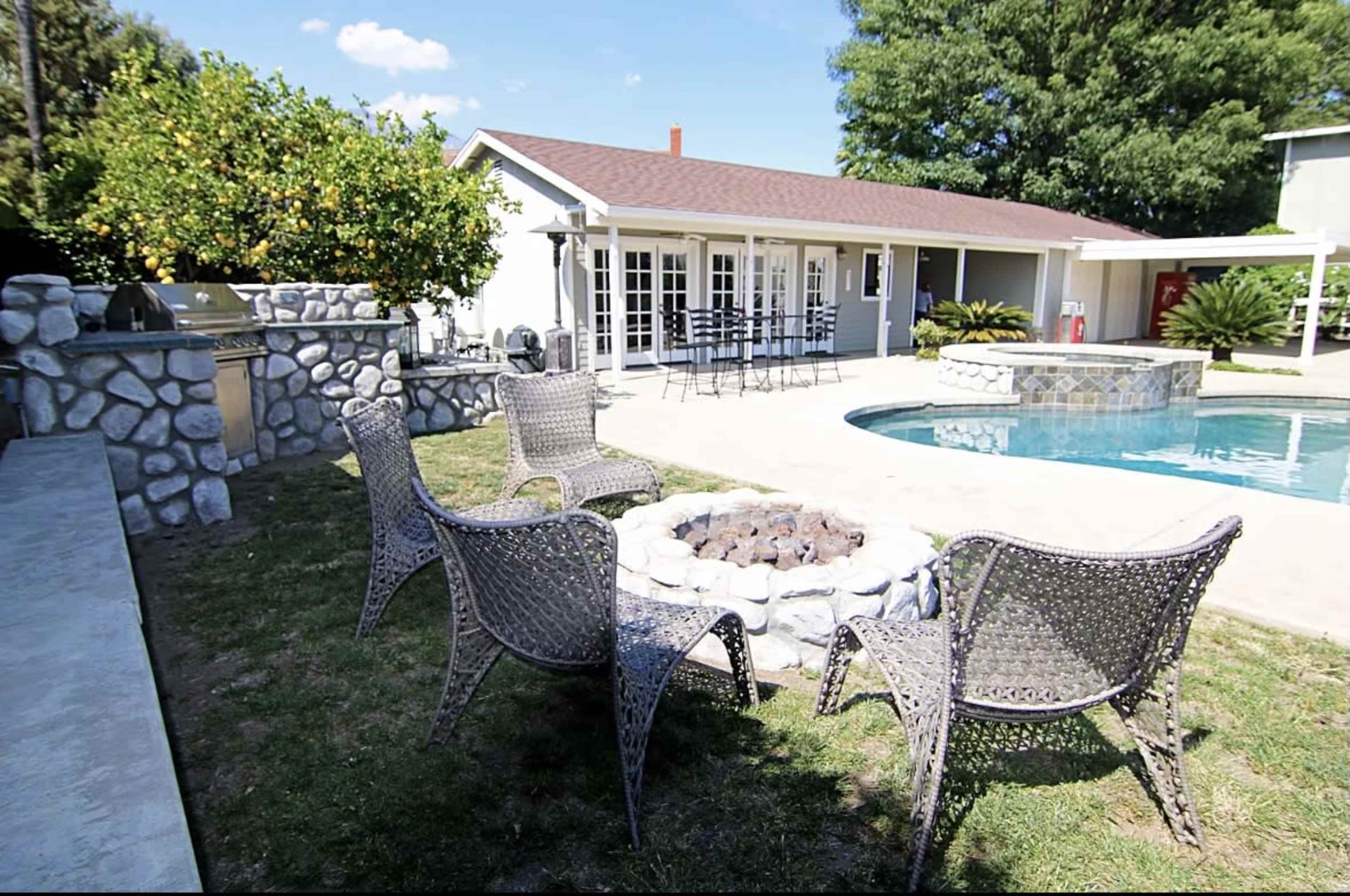A backyard features a swimming pool, a fire pit surrounded by wicker chairs, and a house with a stone barbecue area and outdoor seating.