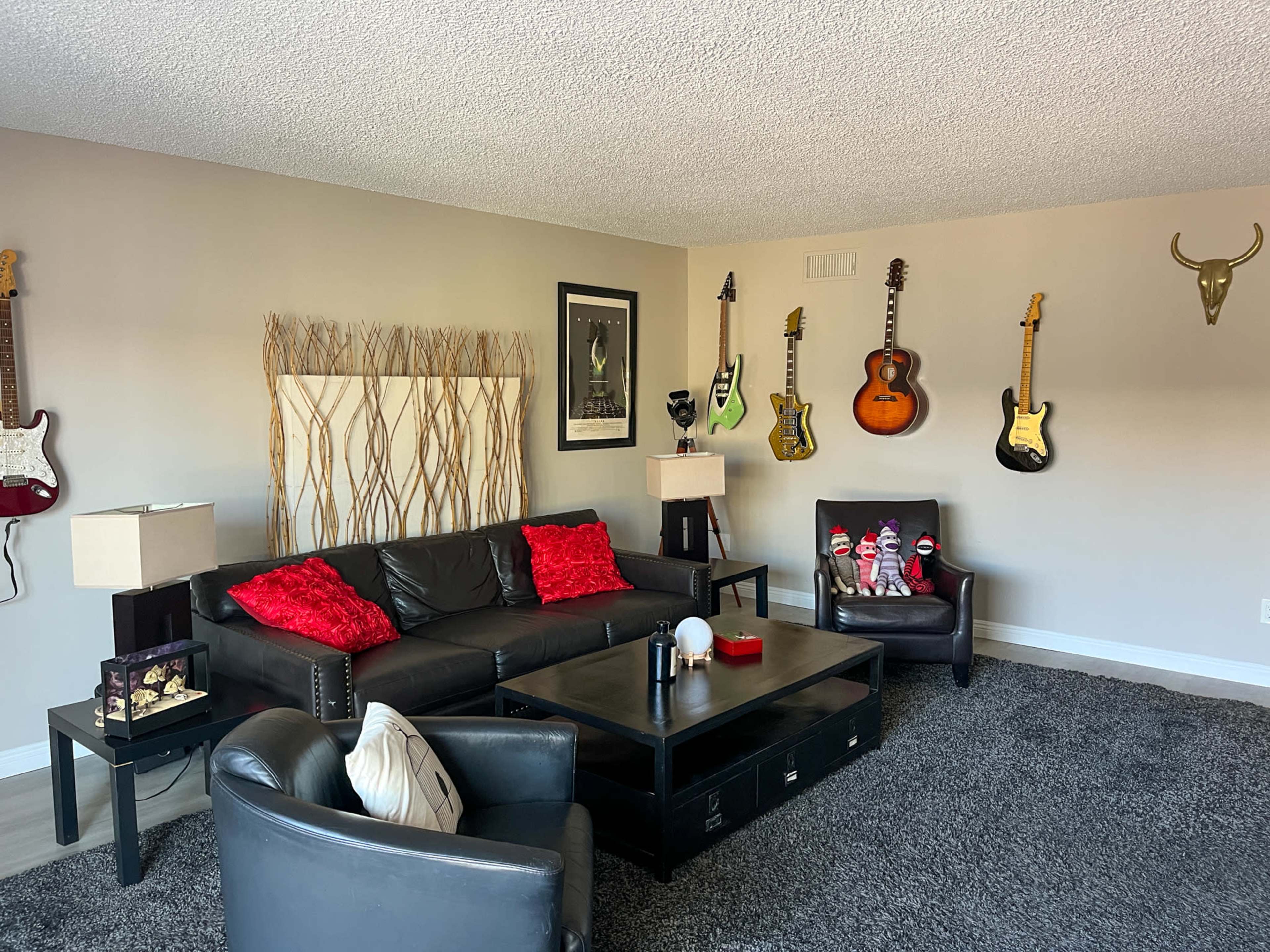 The living room features a black leather sofa, two armchairs, a coffee table, and guitars mounted on the wall, with red decorative pillows and a textured backdrop.