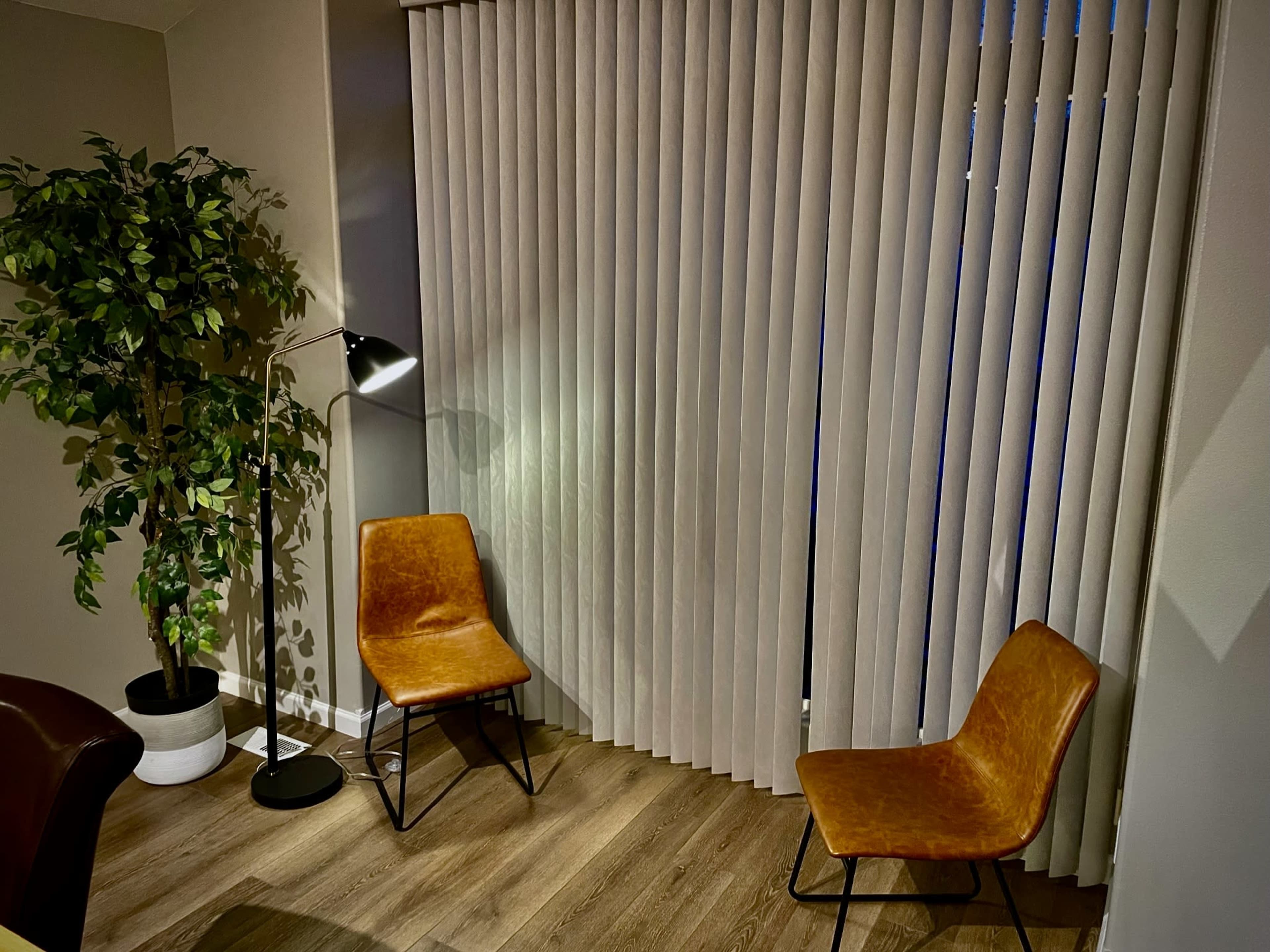 A corner of a room with two brown chairs, a floor lamp, and a potted plant next to vertically striped window coverings.