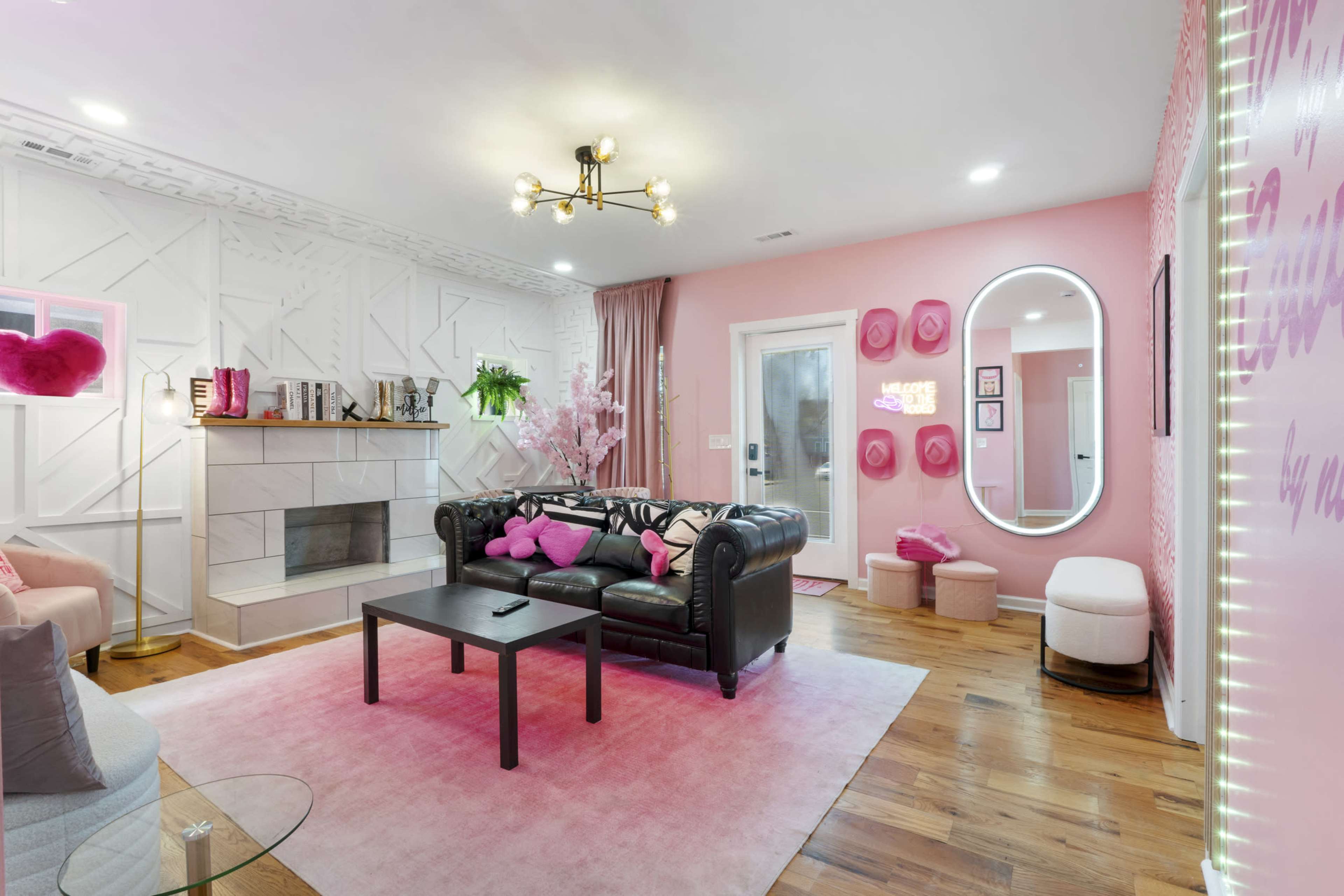The image shows a brightly lit living room featuring pink walls, a black leather sofa, a glass coffee table, and decorative elements such as a fireplace and mirrors.