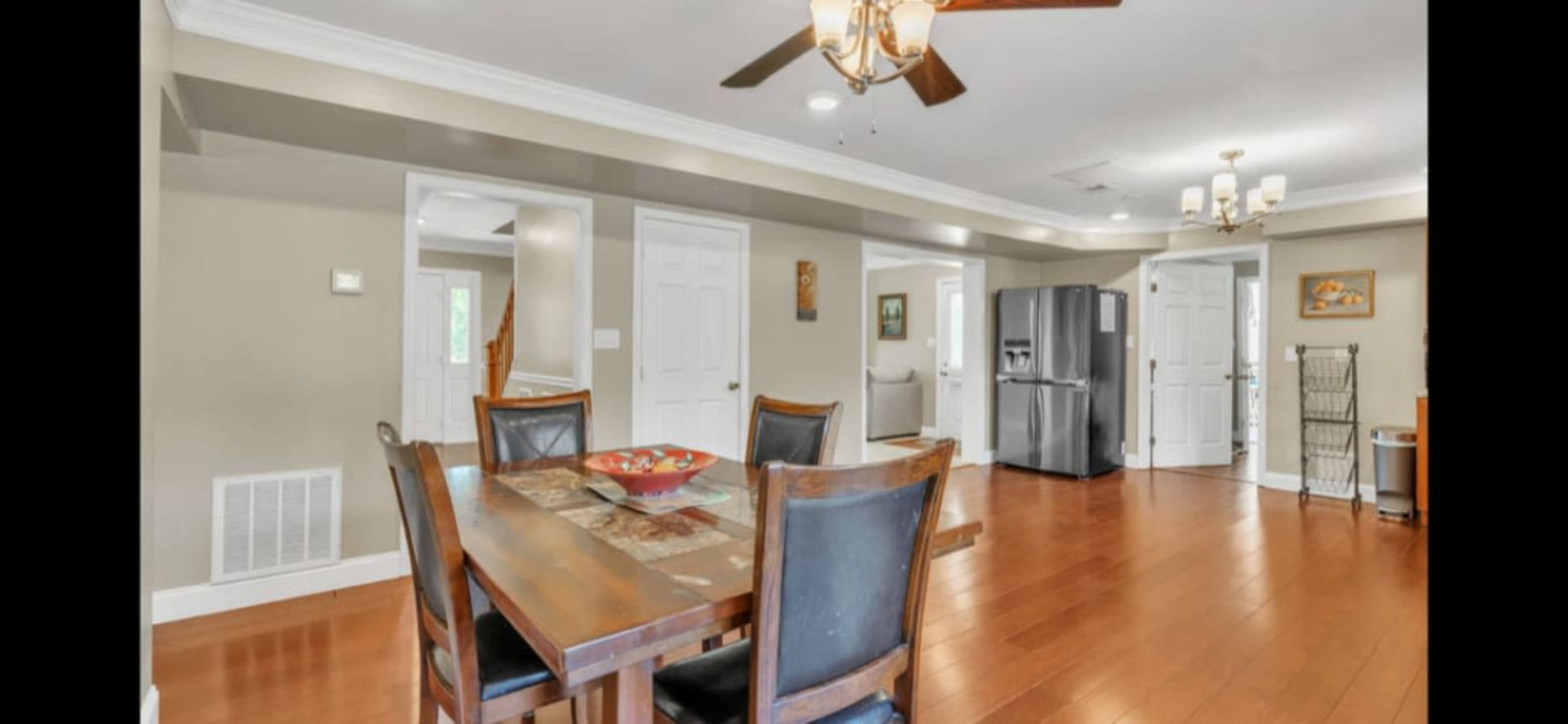 A dining area features a wooden table with chairs, a ceiling fan, and a view of an open kitchen and hallway.