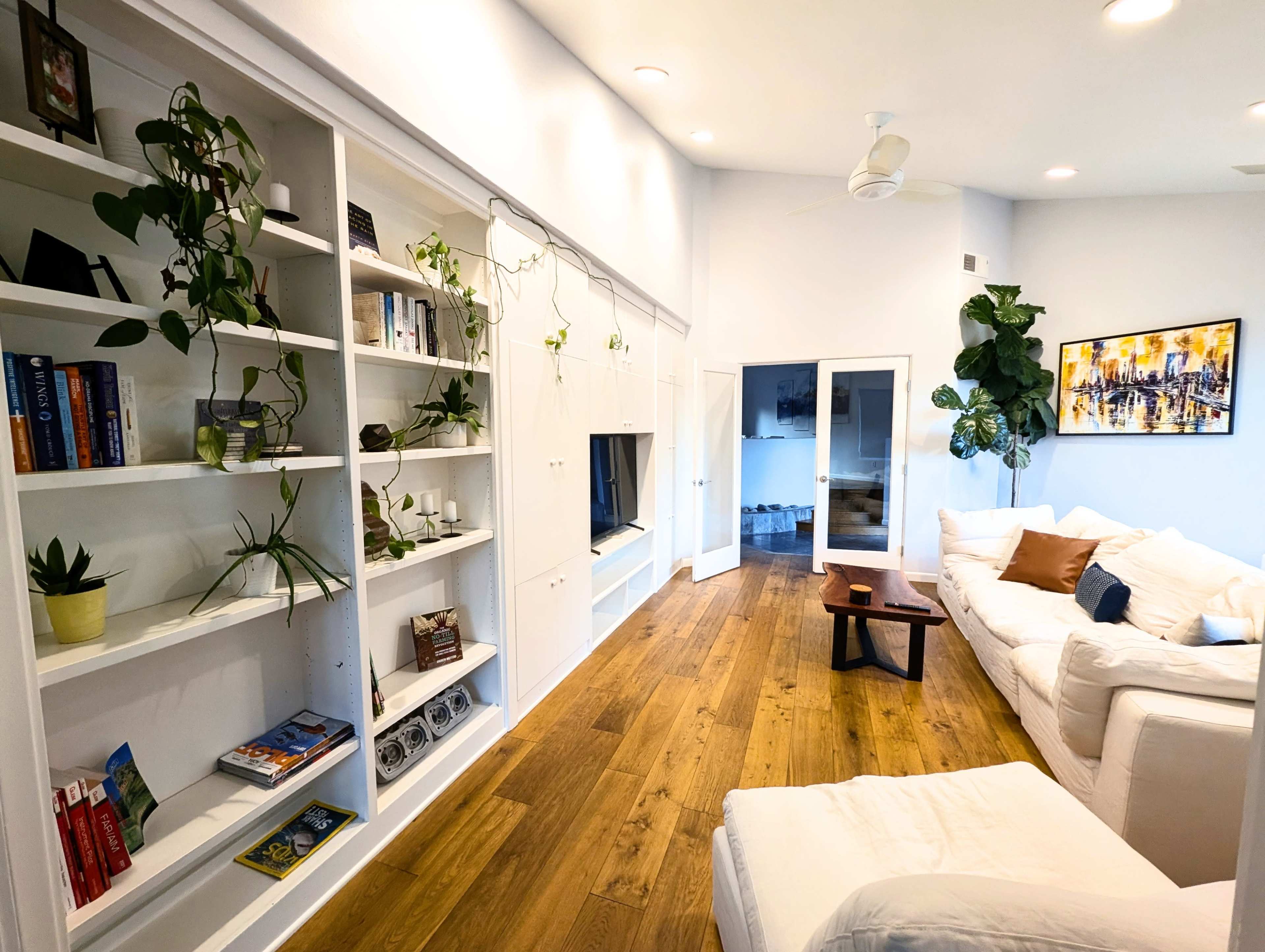 The image depicts a bright, modern living room featuring a white sofa, a wooden coffee table, a bookshelf filled with books and plants, and a large window.