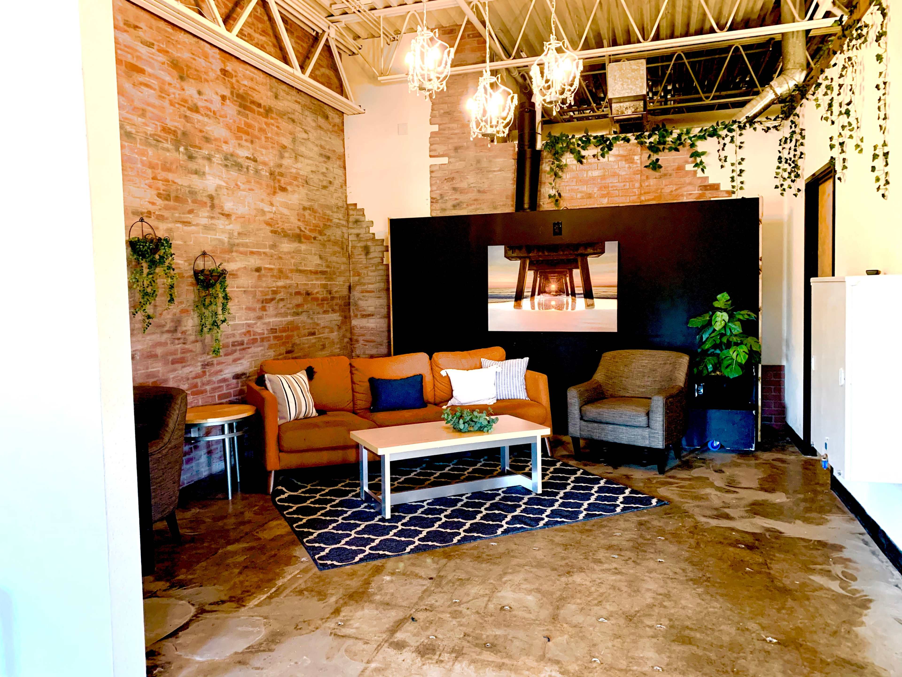 The image shows a modern living space featuring a brown couch, two armchairs, a coffee table, and decorative plants against a brick wall backdrop.