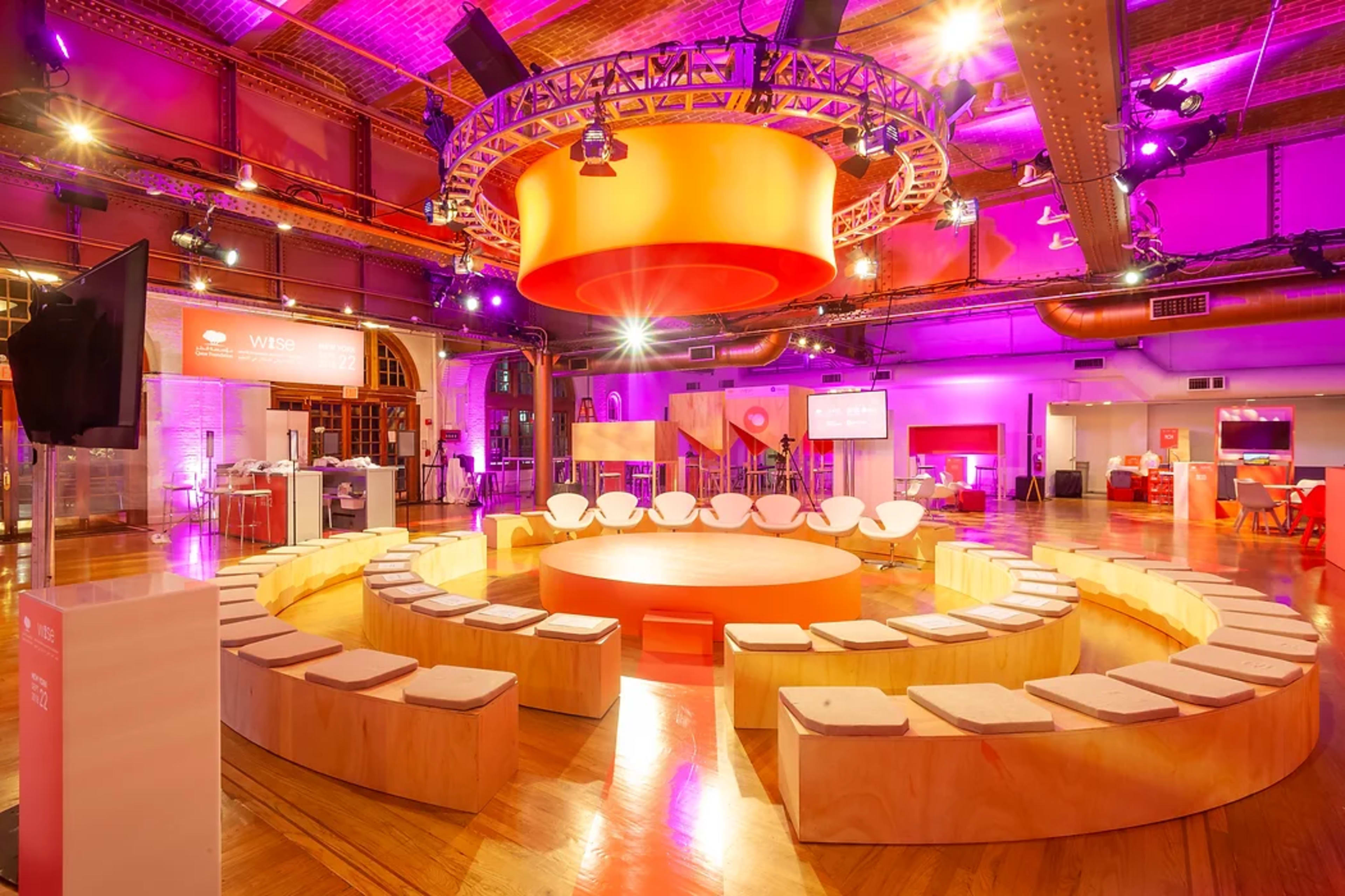 A circular seating arrangement with wooden benches surrounding a central circular platform in a brightly lit event space featuring purple and orange decorations.
