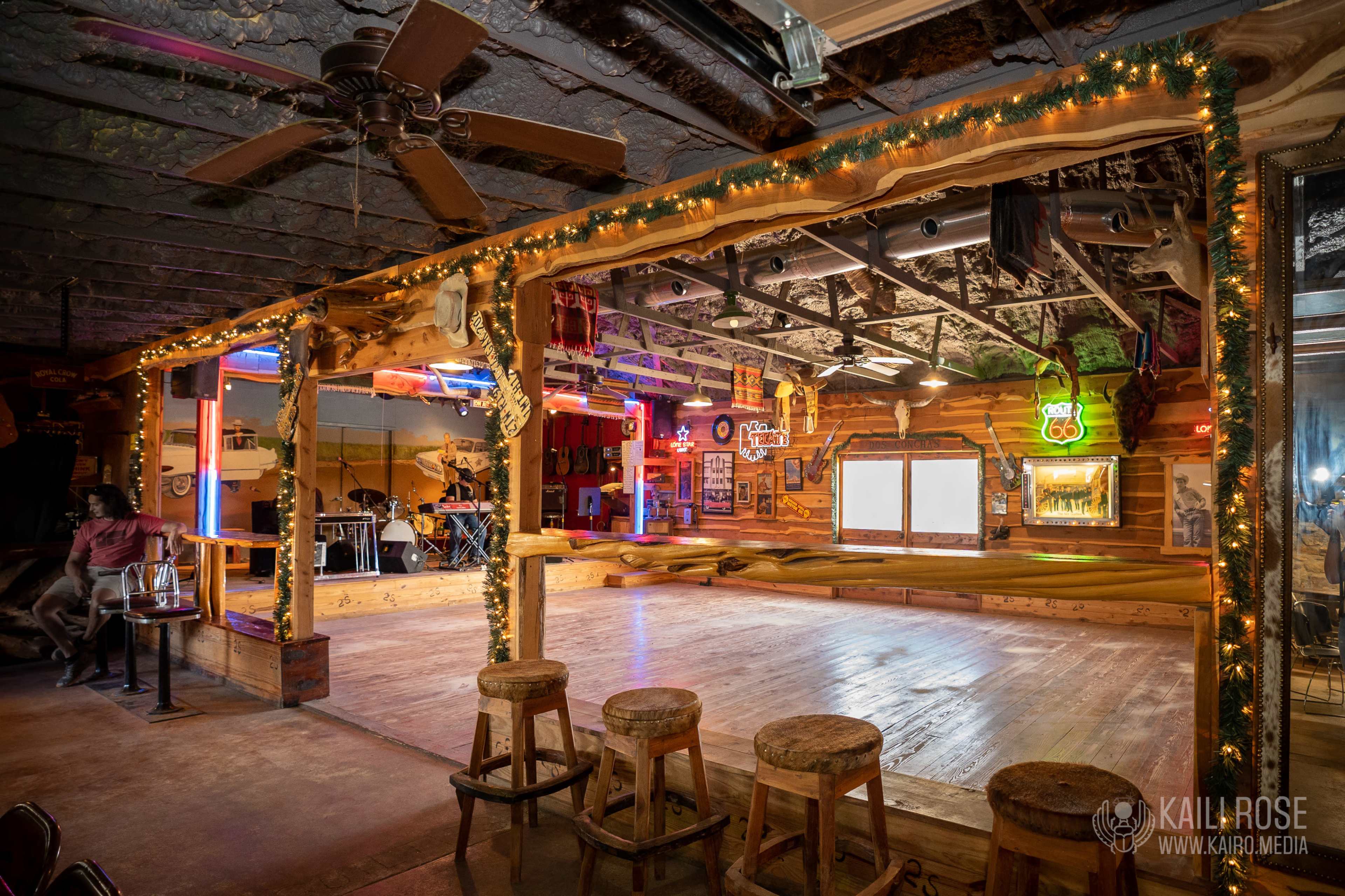 A rustic dance floor area is illuminated by colorful neon lights, with wooden stools and a stage for a band in the background.