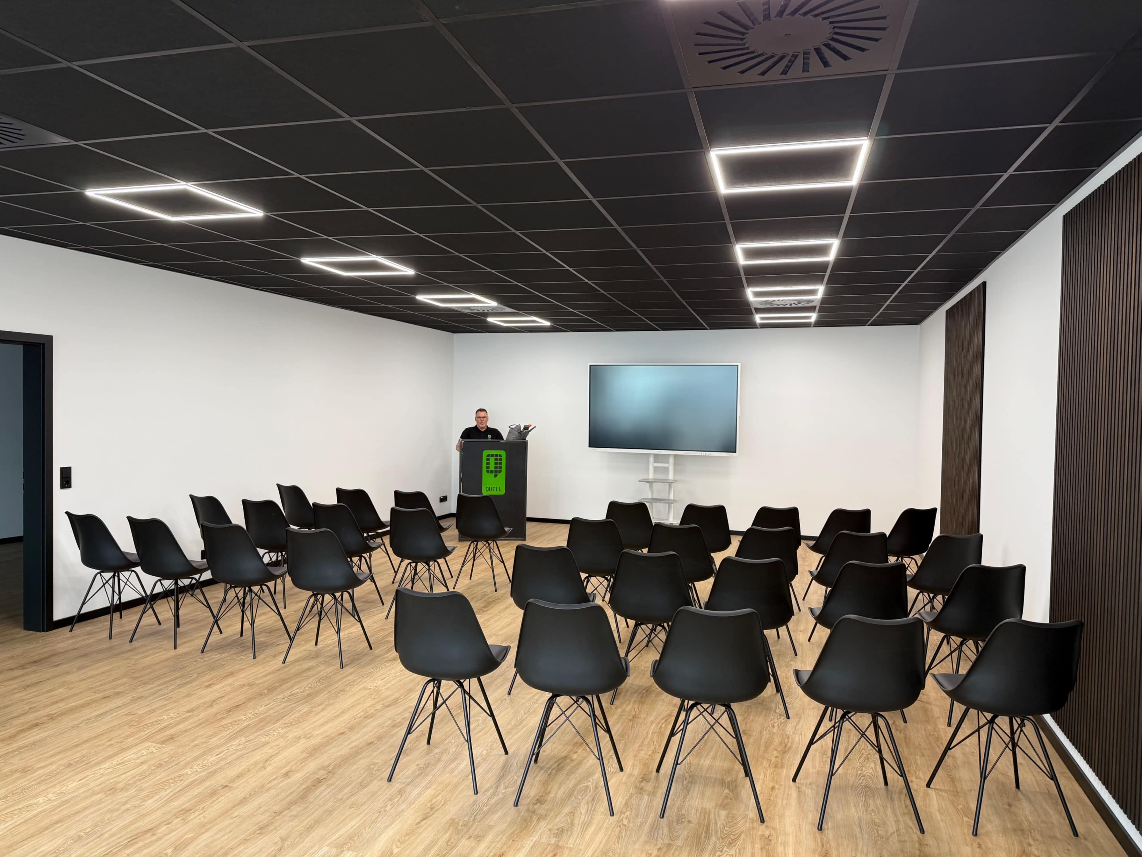 The image shows a conference room with multiple rows of black chairs facing a podium and a wall-mounted screen.