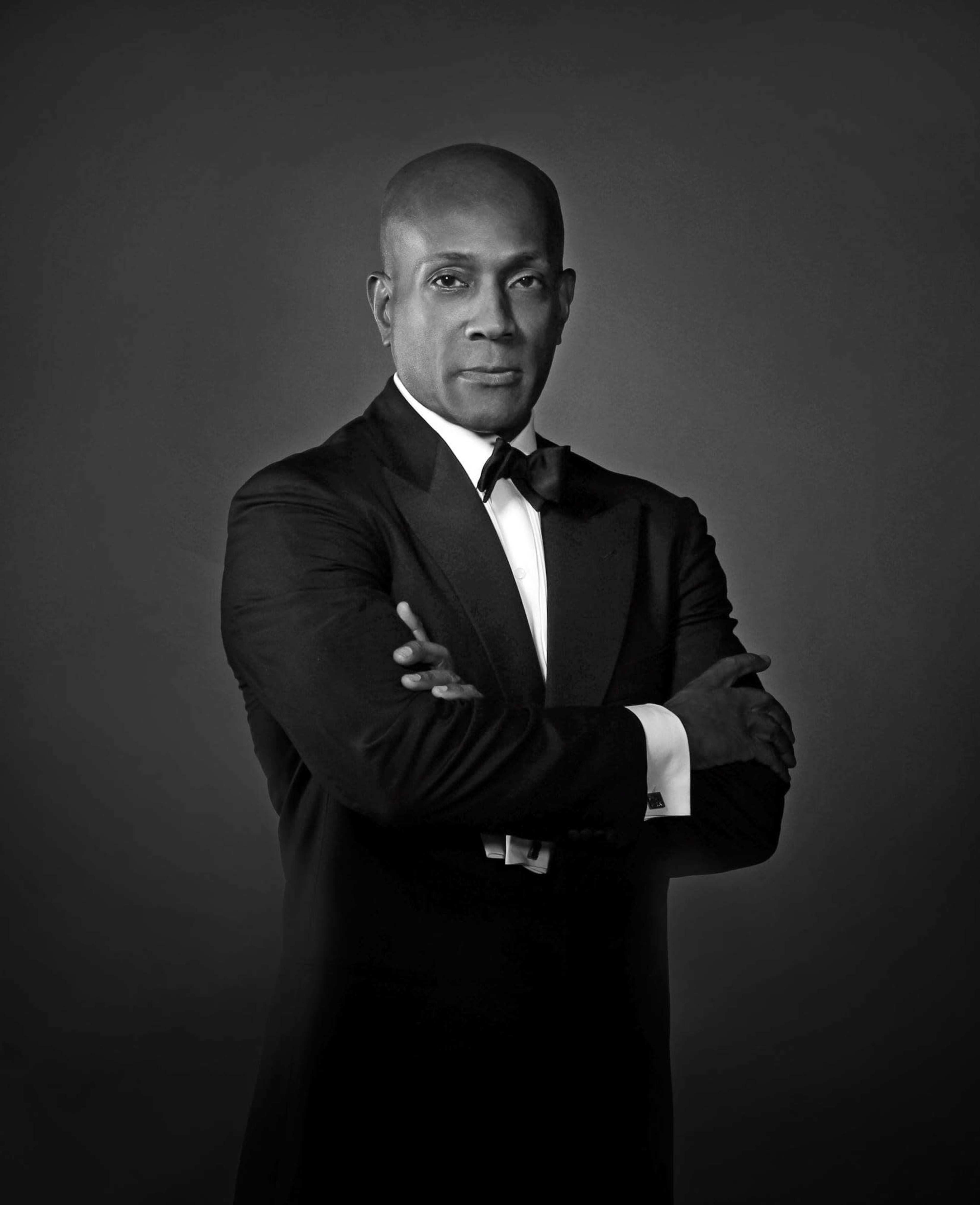 A man in formal attire stands with his arms crossed against a dark background.