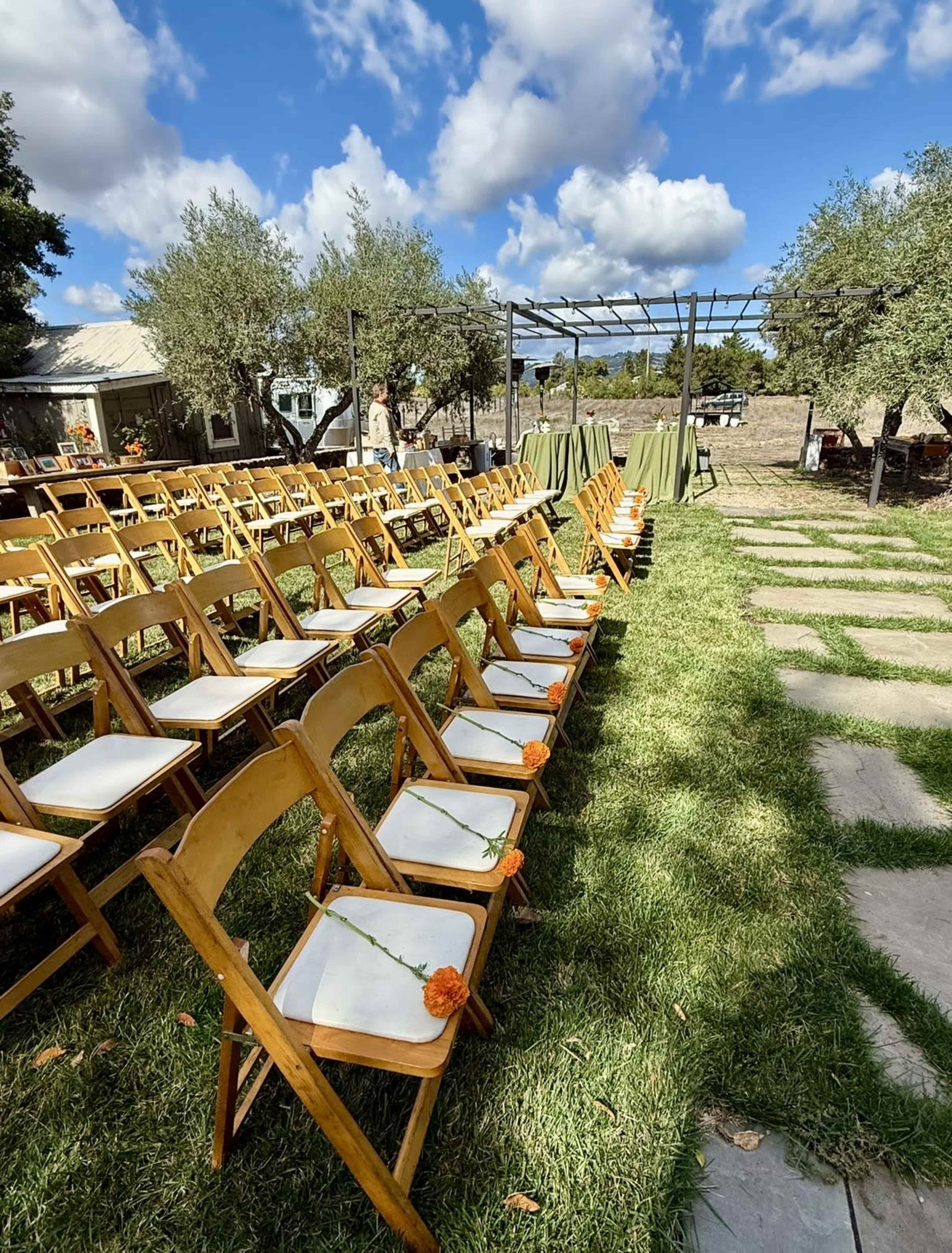 A row of wooden chairs adorned with orange flowers is set up on a grassy area for an outdoor event, under a partly cloudy sky.