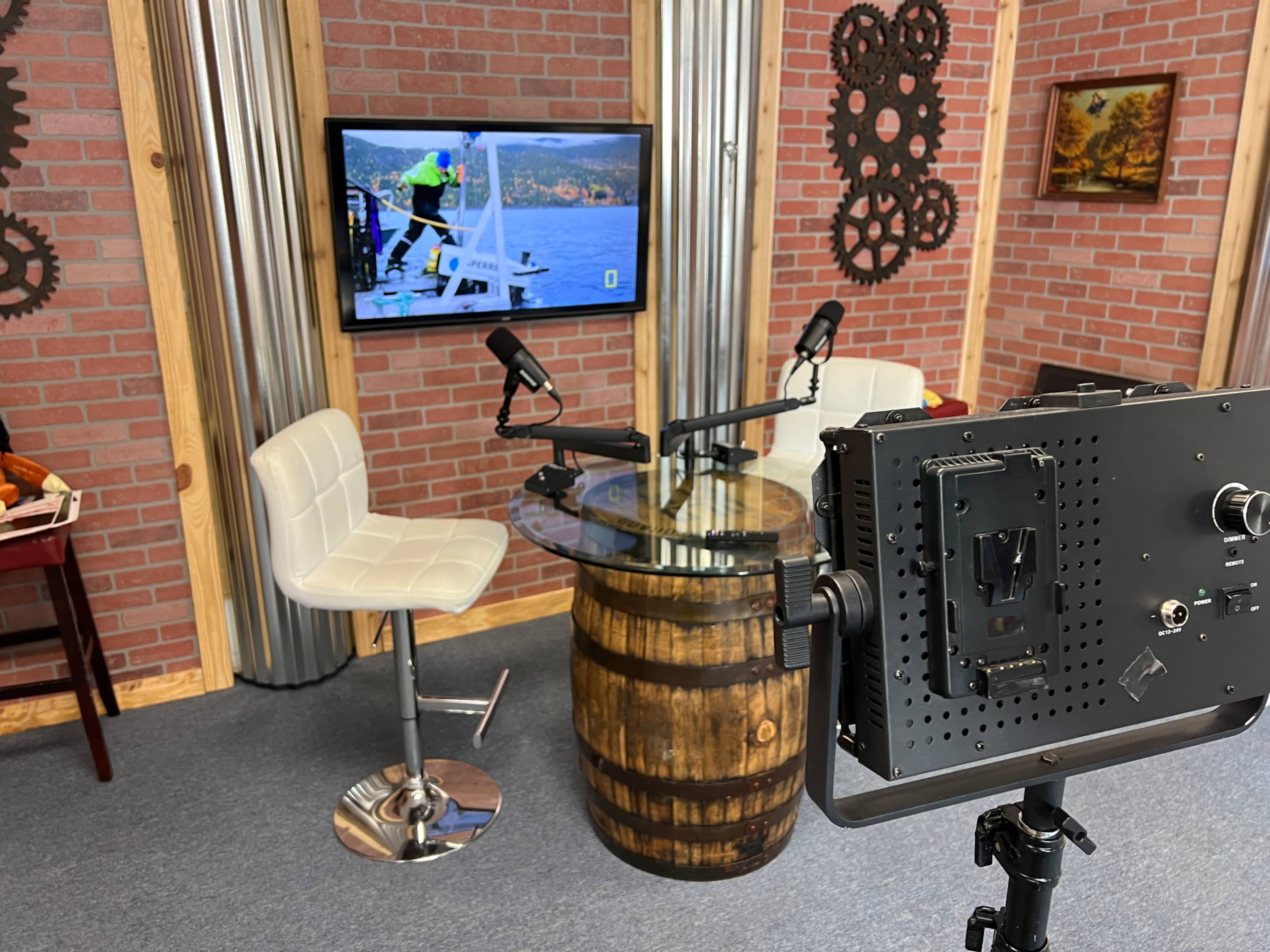 The scene shows a small studio set-up featuring a barrel table, two white chairs, and a television displaying a mountainous landscape, with professional microphone setups.