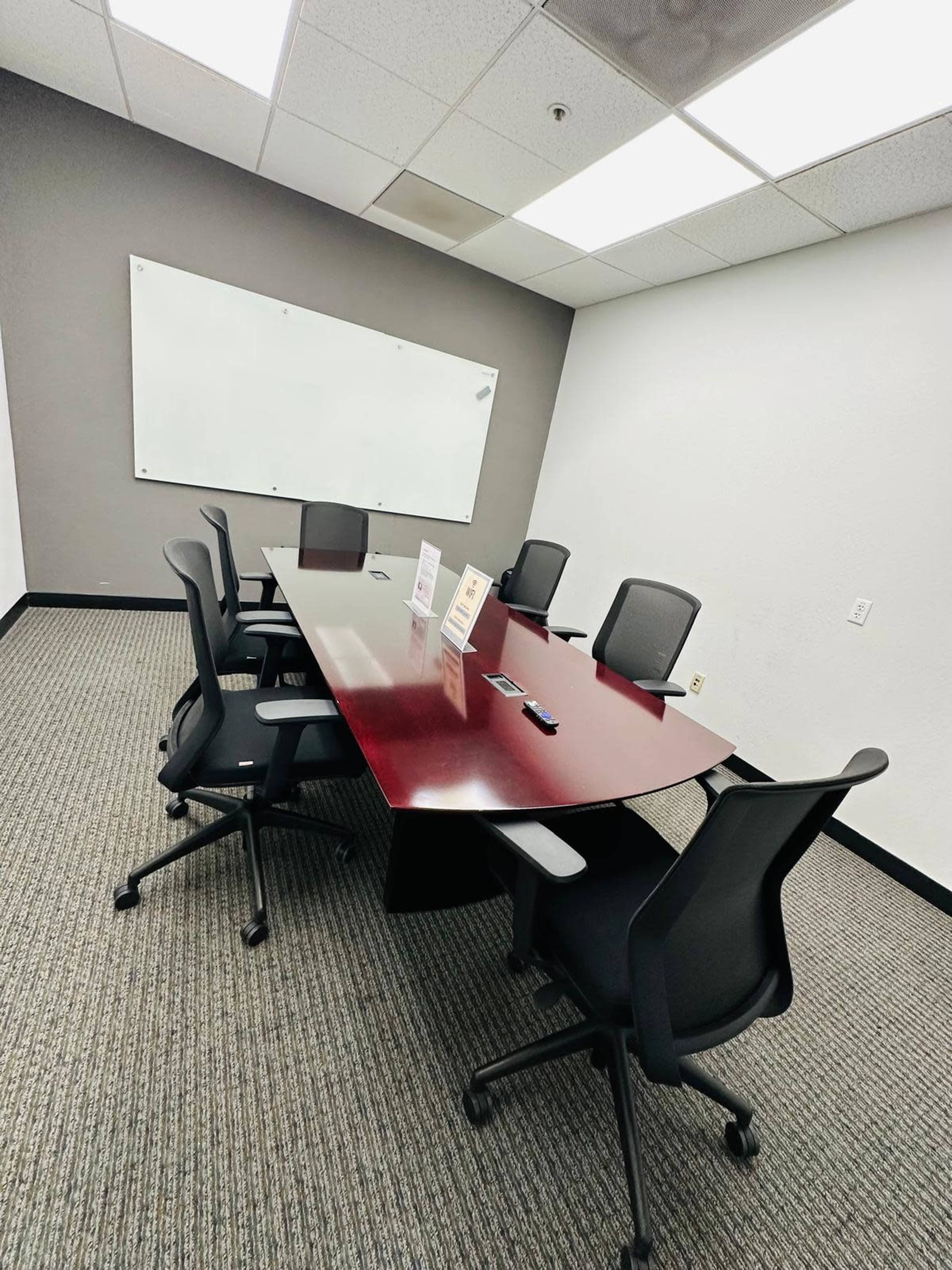 The image shows a conference room with a large wooden table surrounded by six black mesh chairs and a whiteboard on the wall.