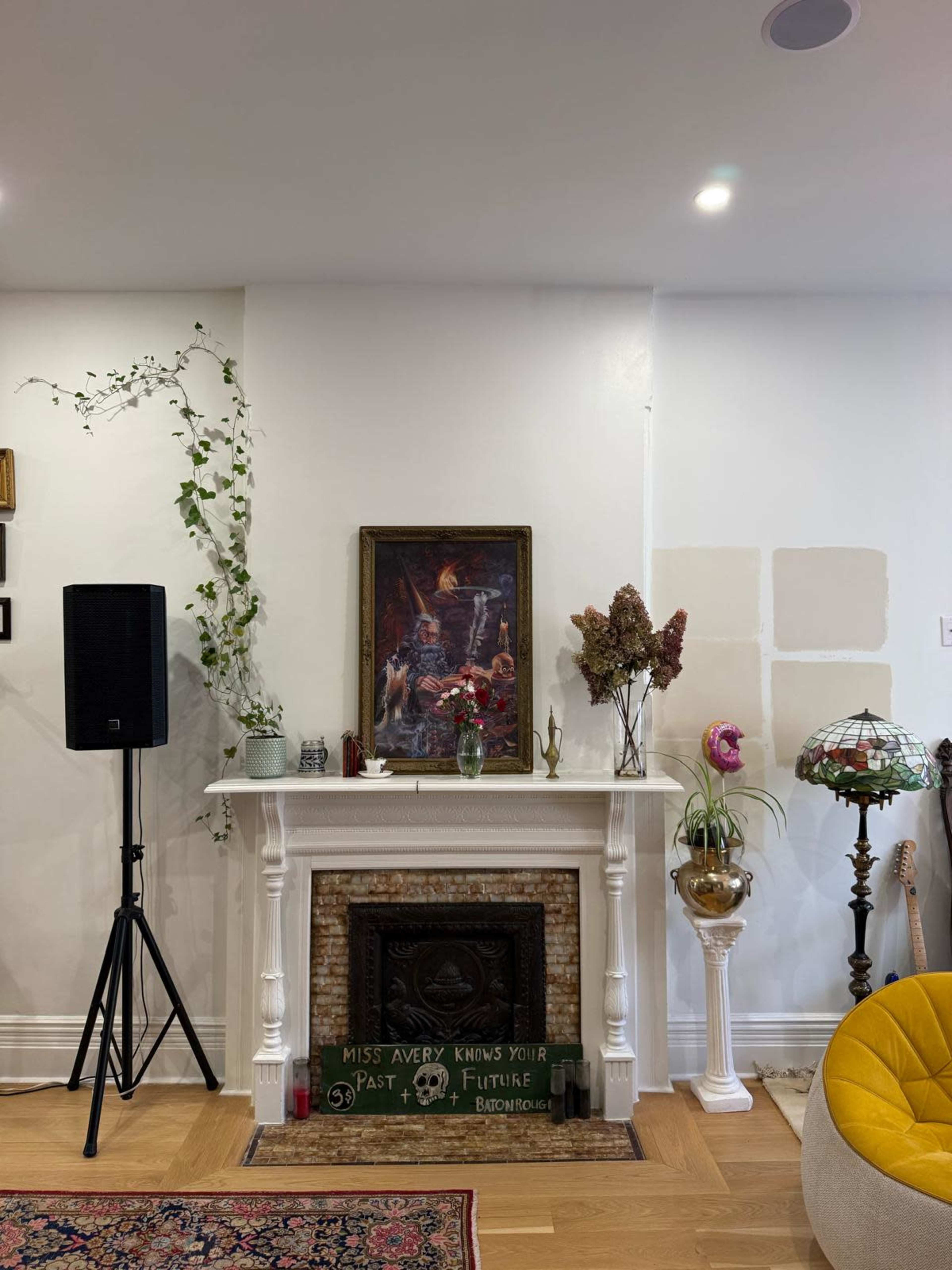 A decorative living room features a fireplace adorned with art, plants, and a vintage lamp, with a speaker and a guitar nearby.