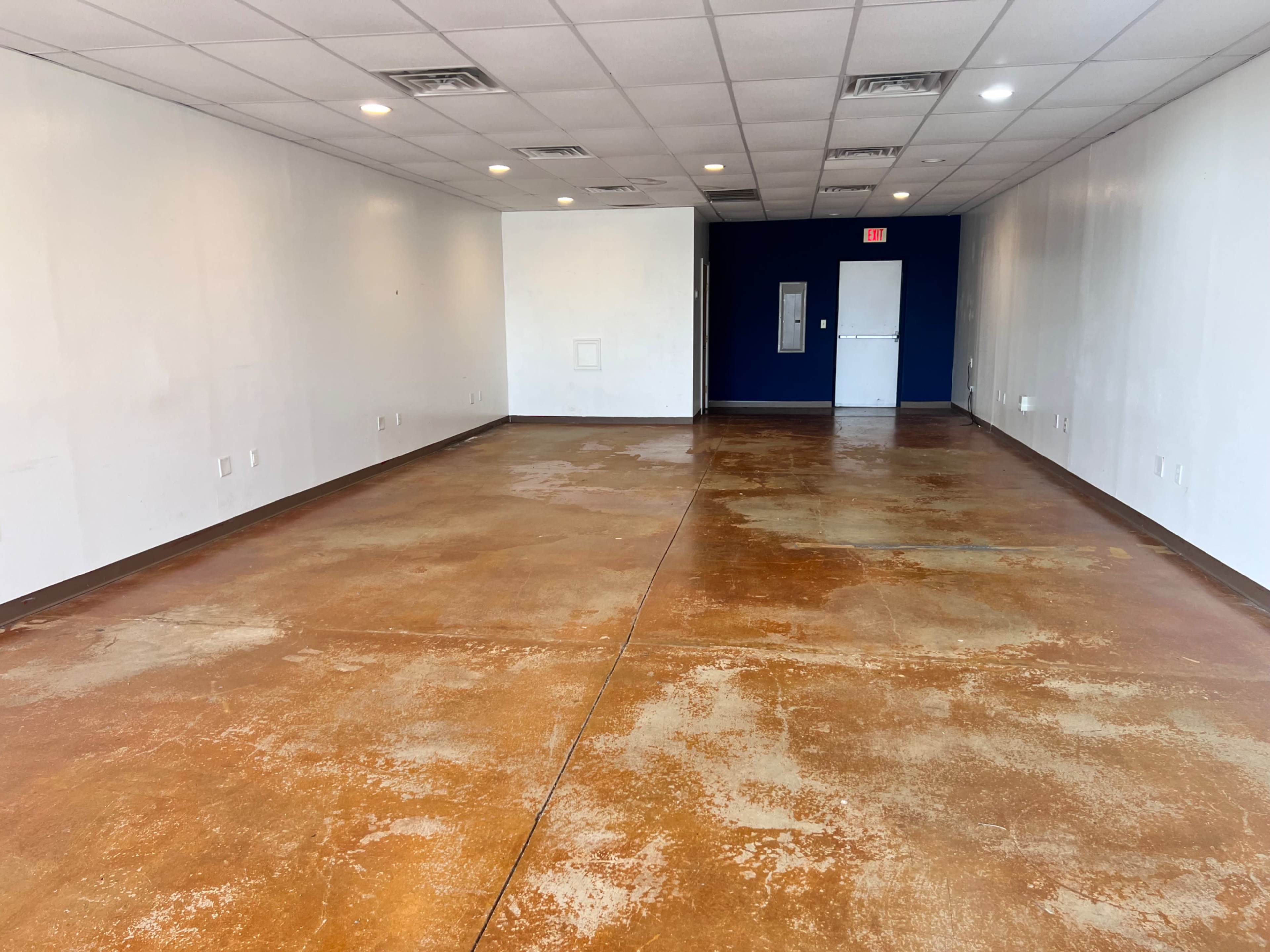The image shows an empty commercial space with a polished concrete floor and white walls, leading to a blue accent wall and a door at the far end.